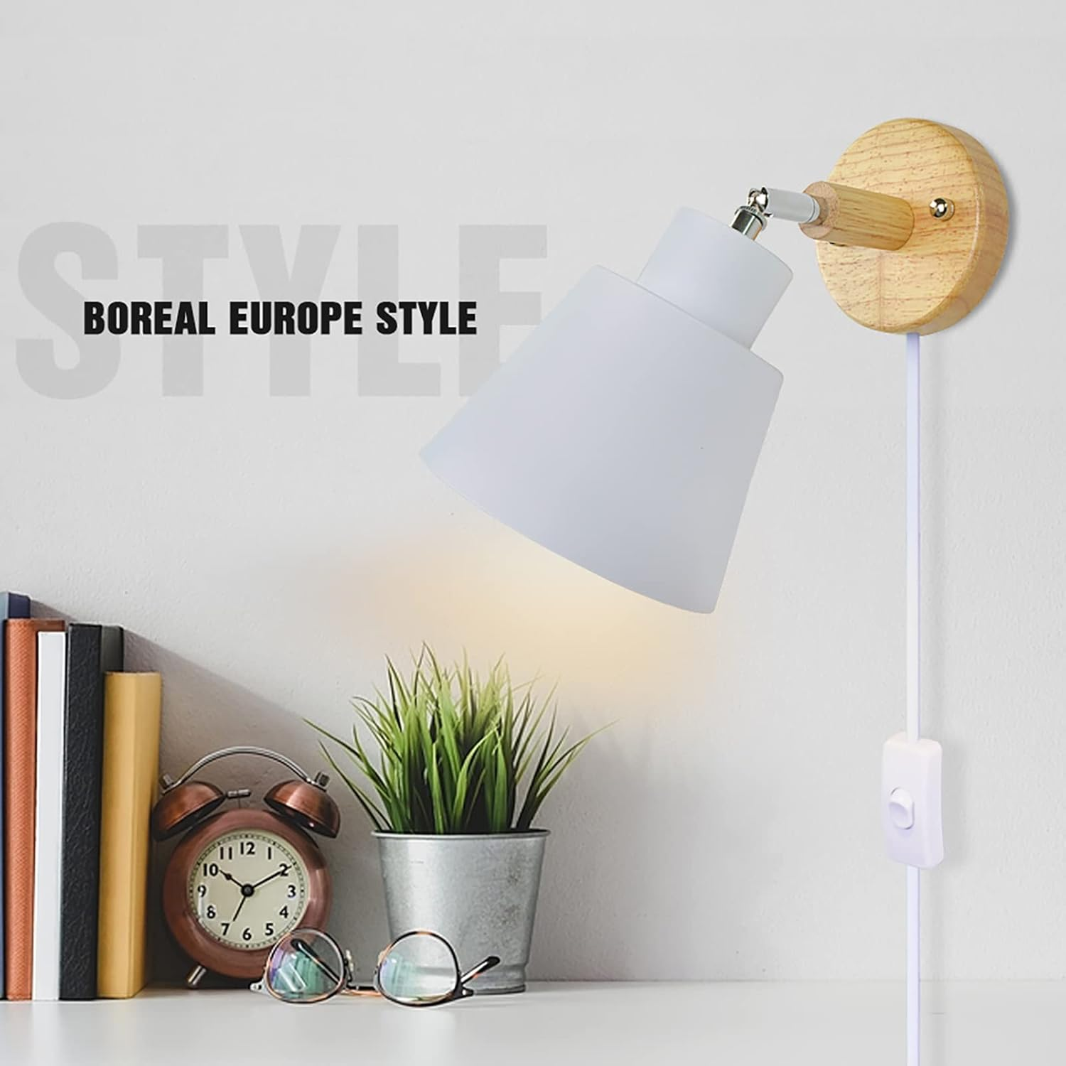 Eine weiße Lampe mit Holzsockel ist an der Wand montiert. Daneben befinden sich eine Uhr, Bücher, eine Pflanze und eine Brille auf einem weißen Regal.