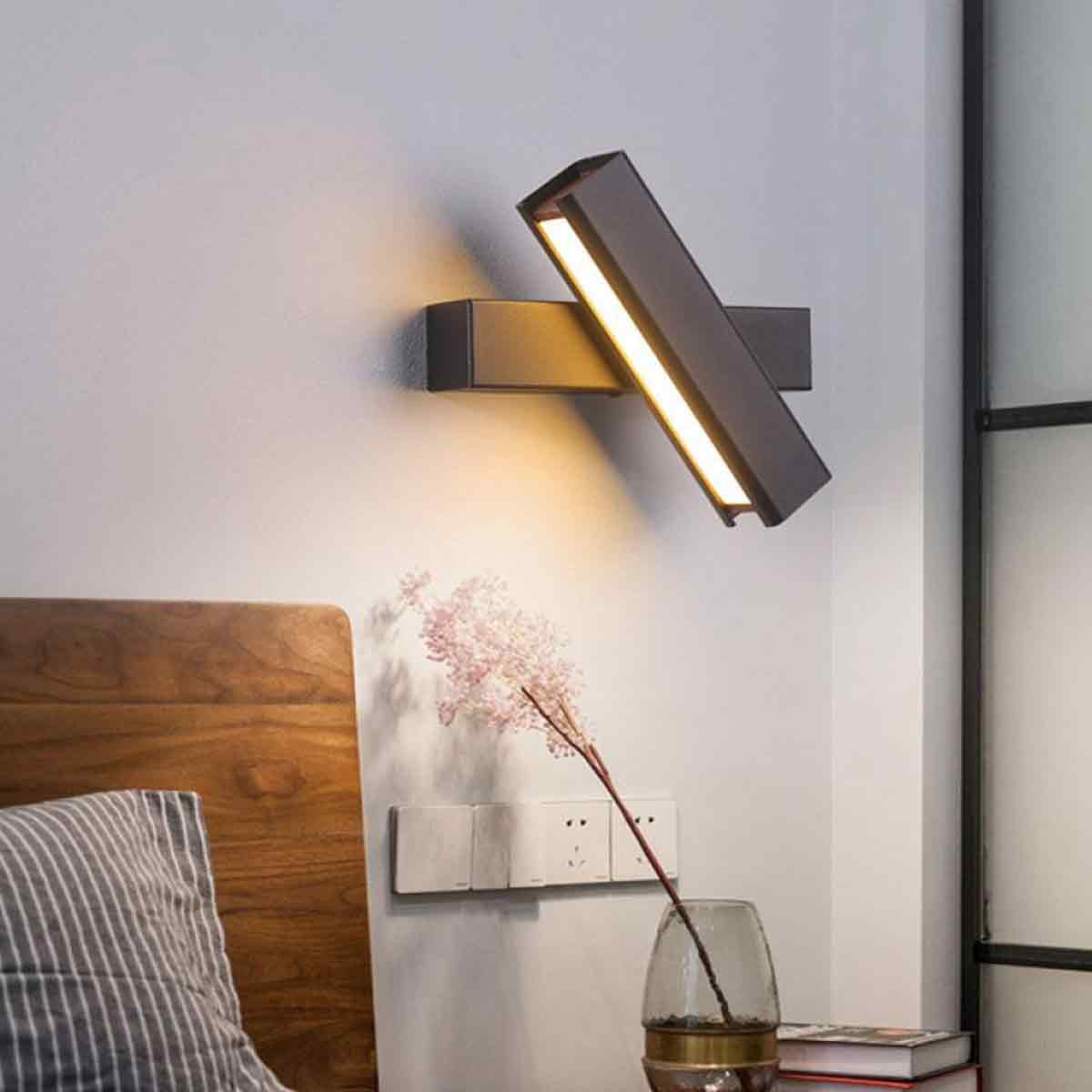 Eine moderne Wandleuchte mit Licht. Es gibt einen Nachttisch mit Vase und Blumen. Ein Bettrahmen und ein Kissen sind im Bild.