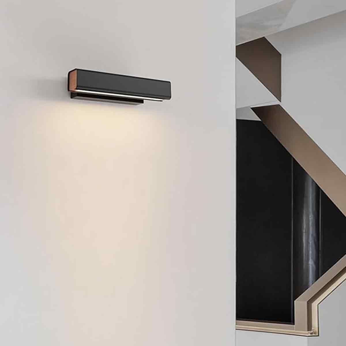 Eine moderne schwarze Wandleuchte strahlt Licht aus. Im Hintergrund befindet sich eine Treppe mit beigen Details. Die Wand ist weiß.
