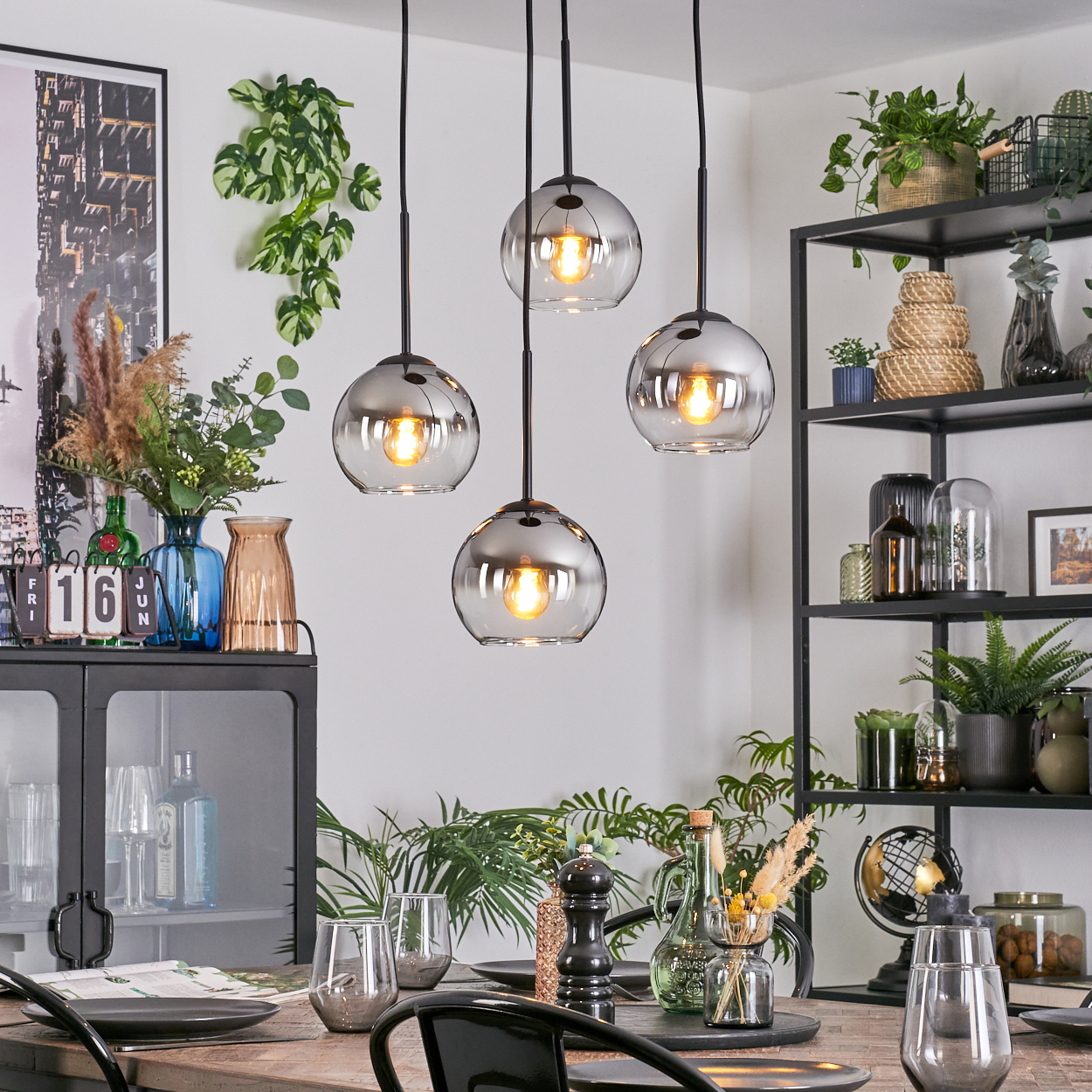 Esszimmer-Szene mit Pendelleuchten, einem gedeckten Tisch und einem schwarzen Metallregal mit Pflanzen und Dekor.