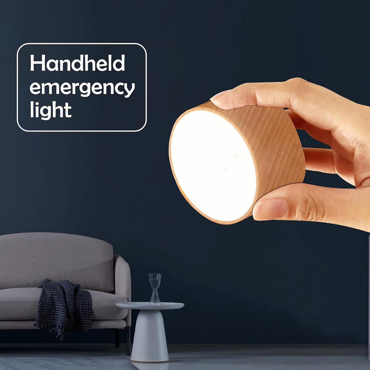 Eine Hand hält eine zylindrische Notfalllampe aus Holz, die helles weißes Licht ausstrahlt. Im Hintergrund sind ein Sofa und ein Tisch.