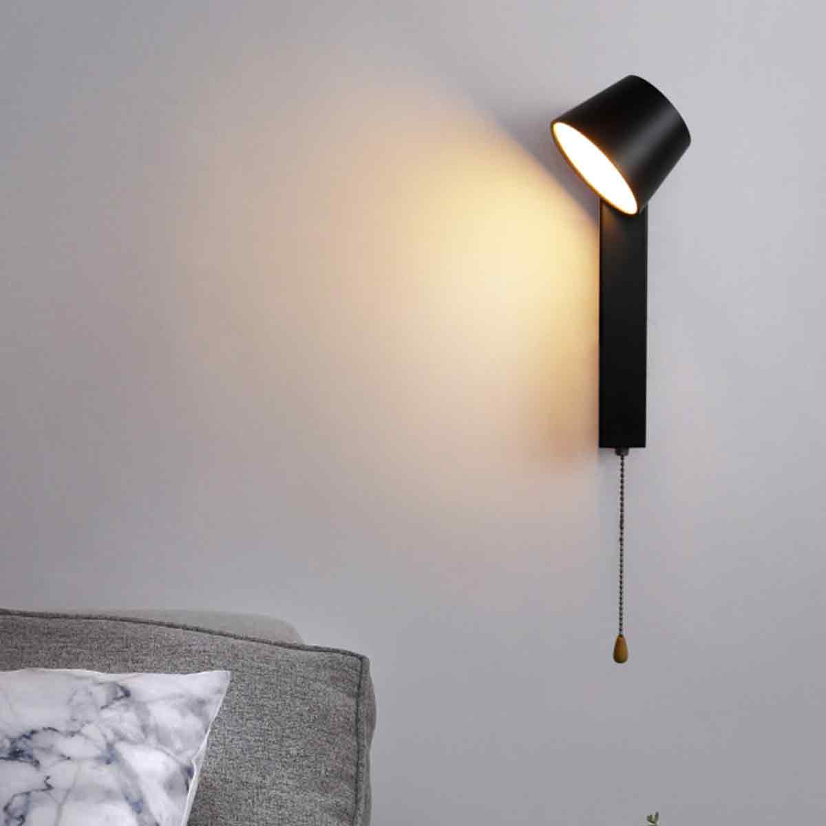 Eine moderne schwarze Wandleuchte strahlt Licht auf eine Wand. Im Vordergrund befinden sich ein Sofa und ein dekoratives Kissen.