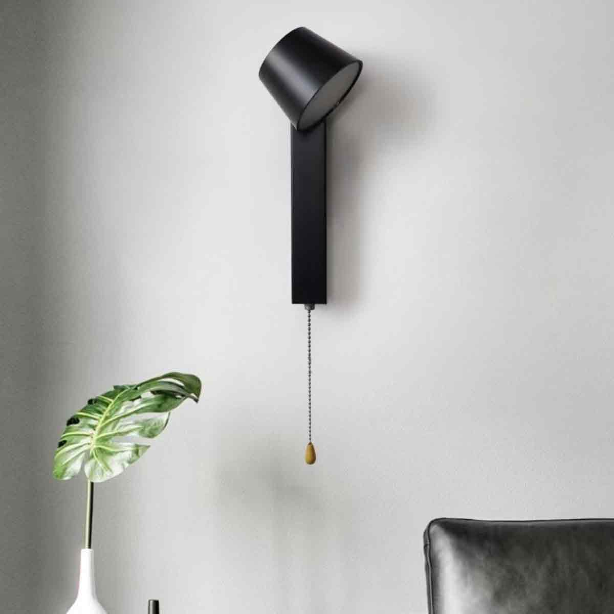 Eine moderne schwarze Wandlampe mit einer Zugkette hängt an einer weißen Wand. Eine Vase mit einer Pflanze ist im Vordergrund.