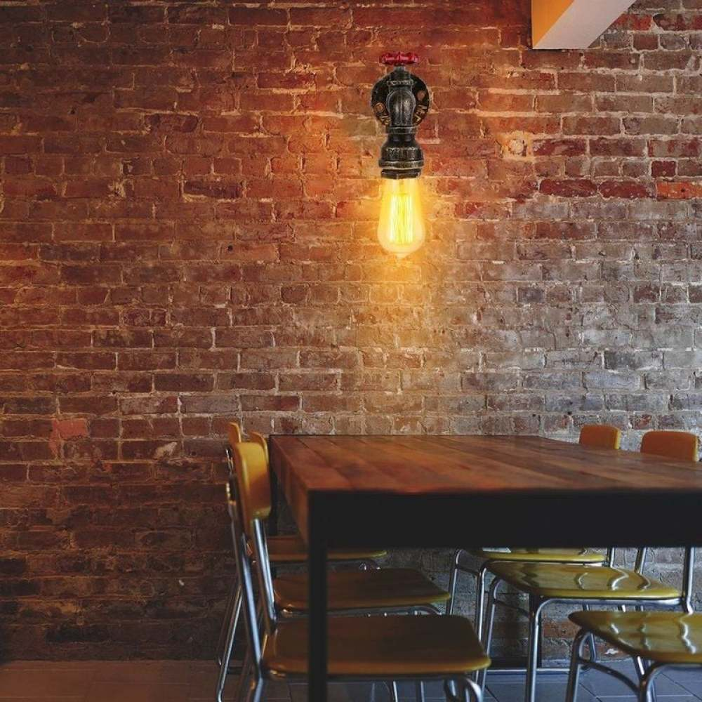 Eine Lampe im Industriestil mit sichtbarer Glühbirne hängt an einer Backsteinmauer. Davor stehen ein Holztisch und gelbe Stühle.