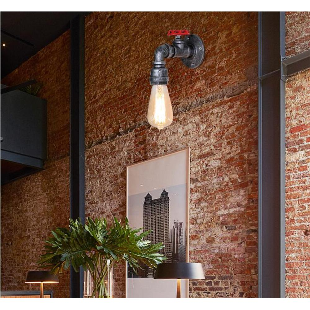 Eine Lampe im Industriestil mit rotem Ventil ist an einer Ziegelmauer angebracht. Eine leuchtende Glühbirne hängt darunter. Ein Bild und Pflanzen sind davor.
