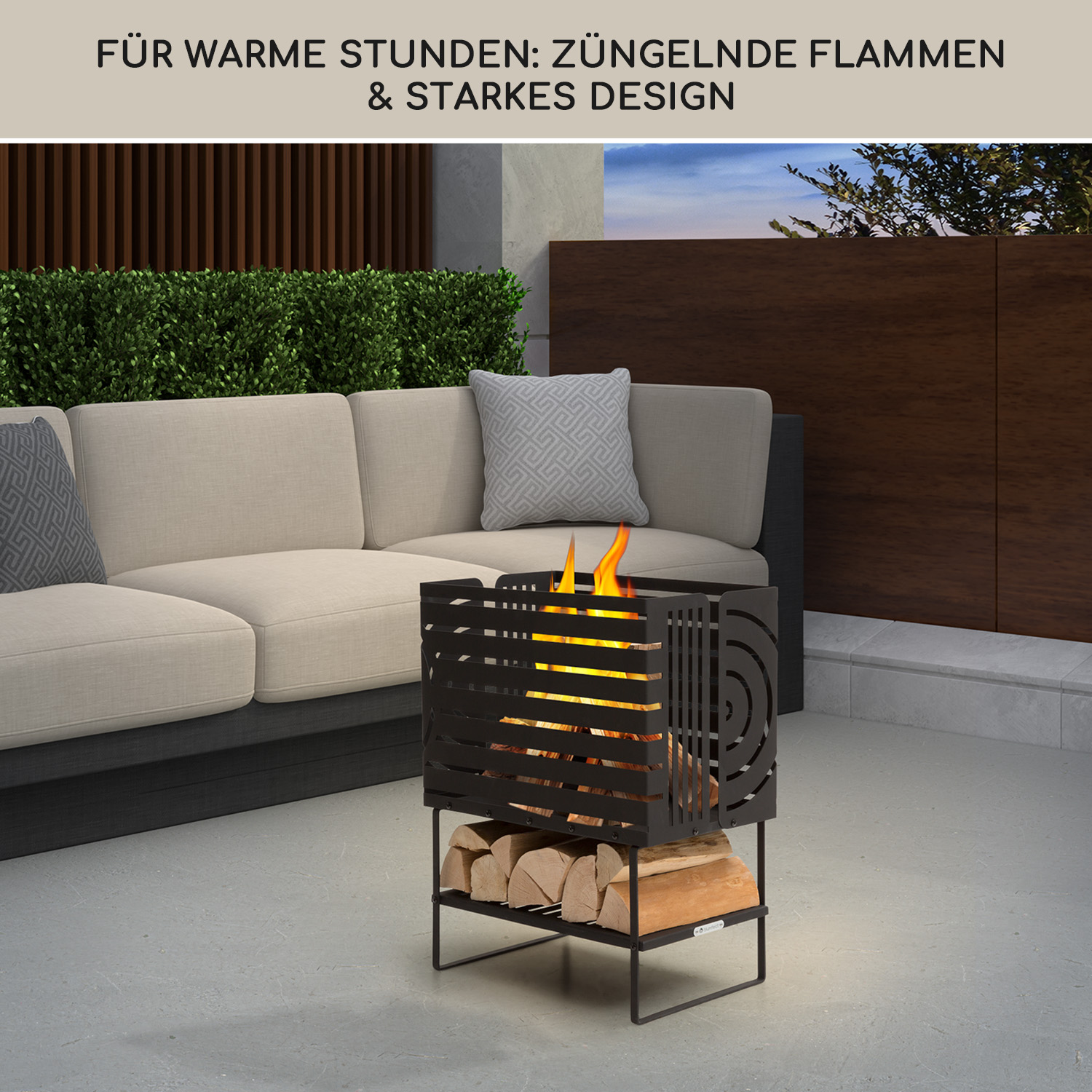 Ein schwarzer Metallfeuerkorb mit brennenden Flammen, Holzscheiten darunter und einem Sofa im Hintergrund auf einer Terrasse.
