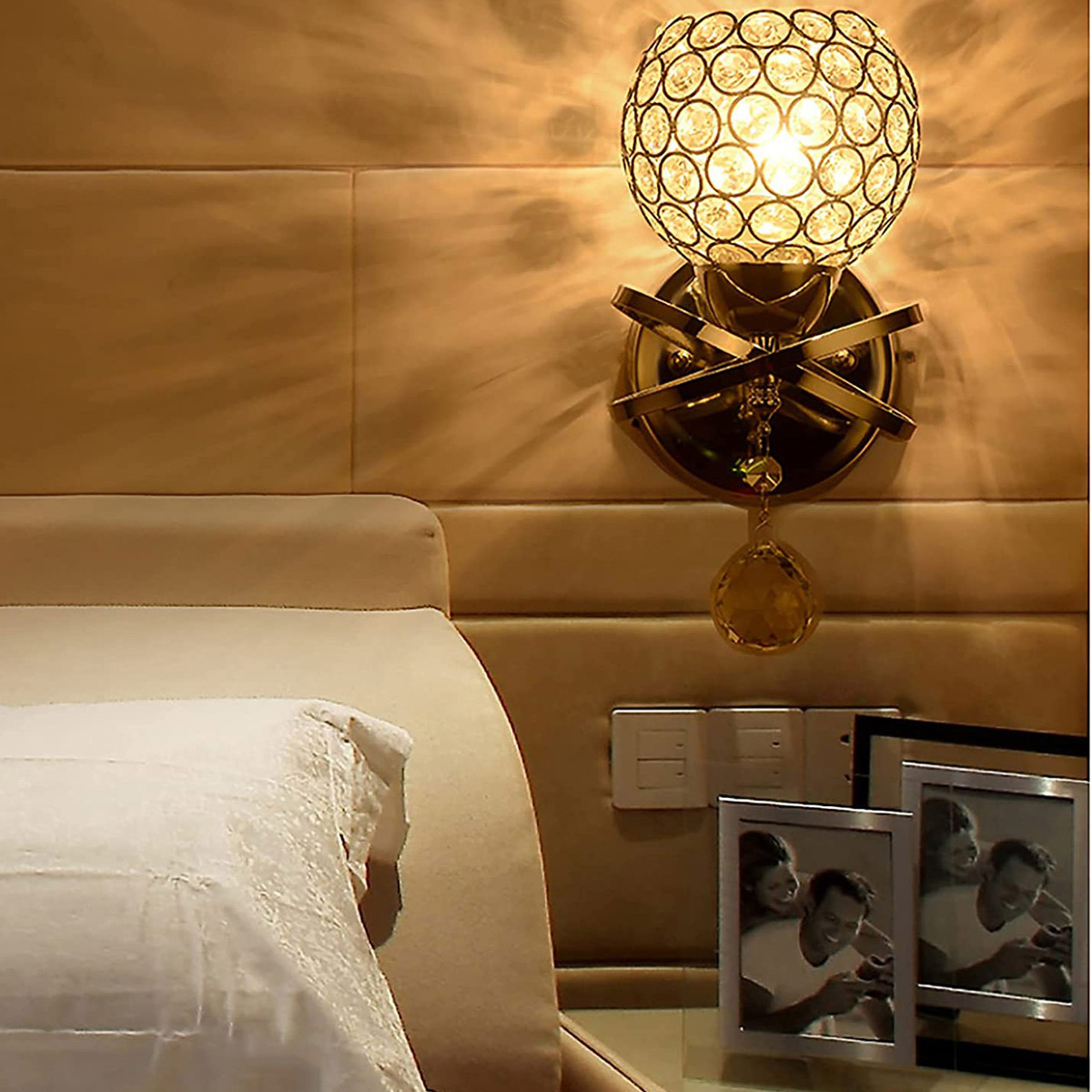 Eine kristallkugelförmige Wandlampe ist an einer gepolsterten Wand befestigt. Ein Bett und gerahmte Fotos sind im Vordergrund.