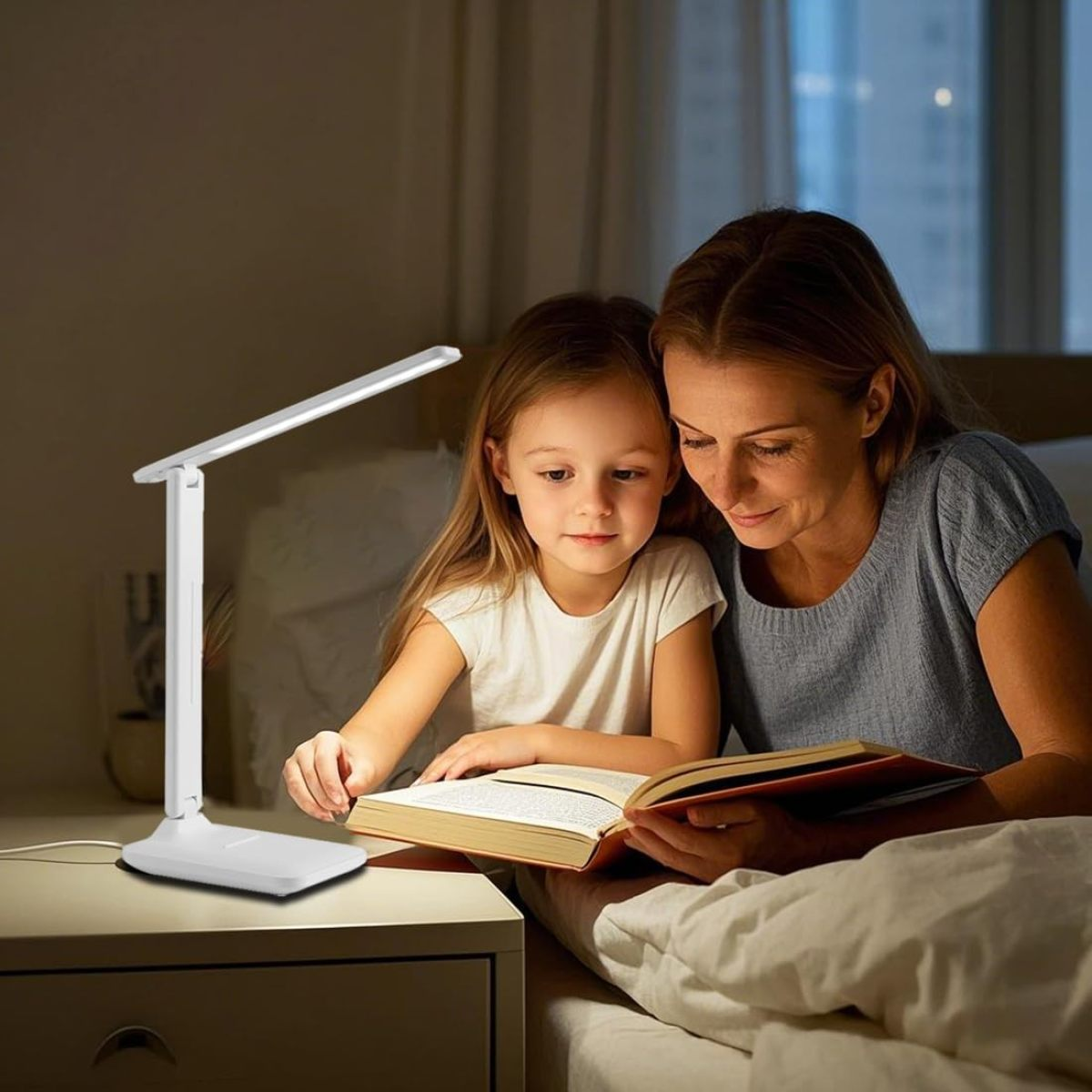 Eine Frau und ein Mädchen lesen unter einer beleuchteten Lampe auf einem Nachttisch ein Buch. Die Szene ist in einem Schlafzimmer, sanftes Licht.