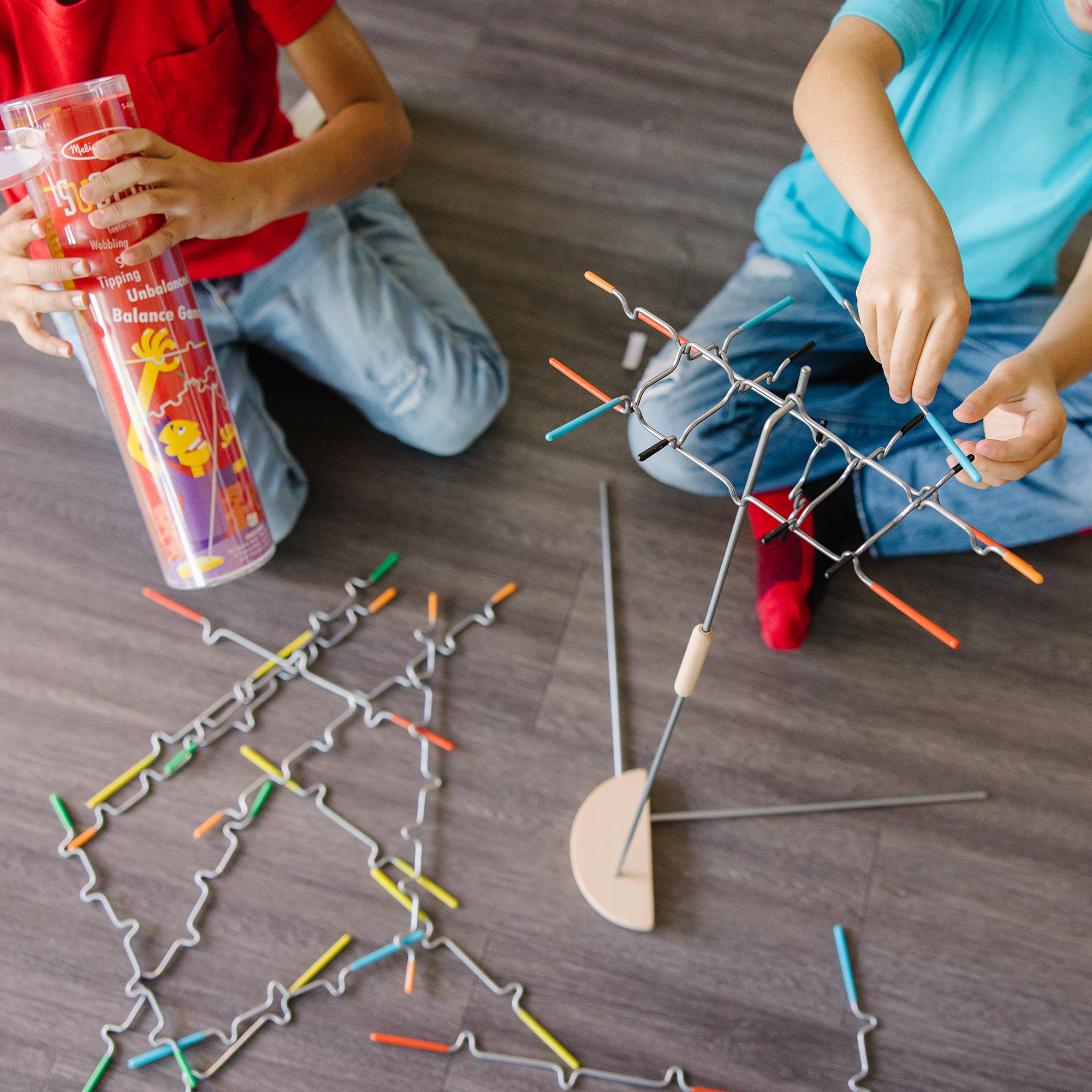 Zwei Kinder spielen mit einem Balance-Spiel auf einem Holzboden. Einer hält die Spielbox, der andere baut die Struktur zusammen.