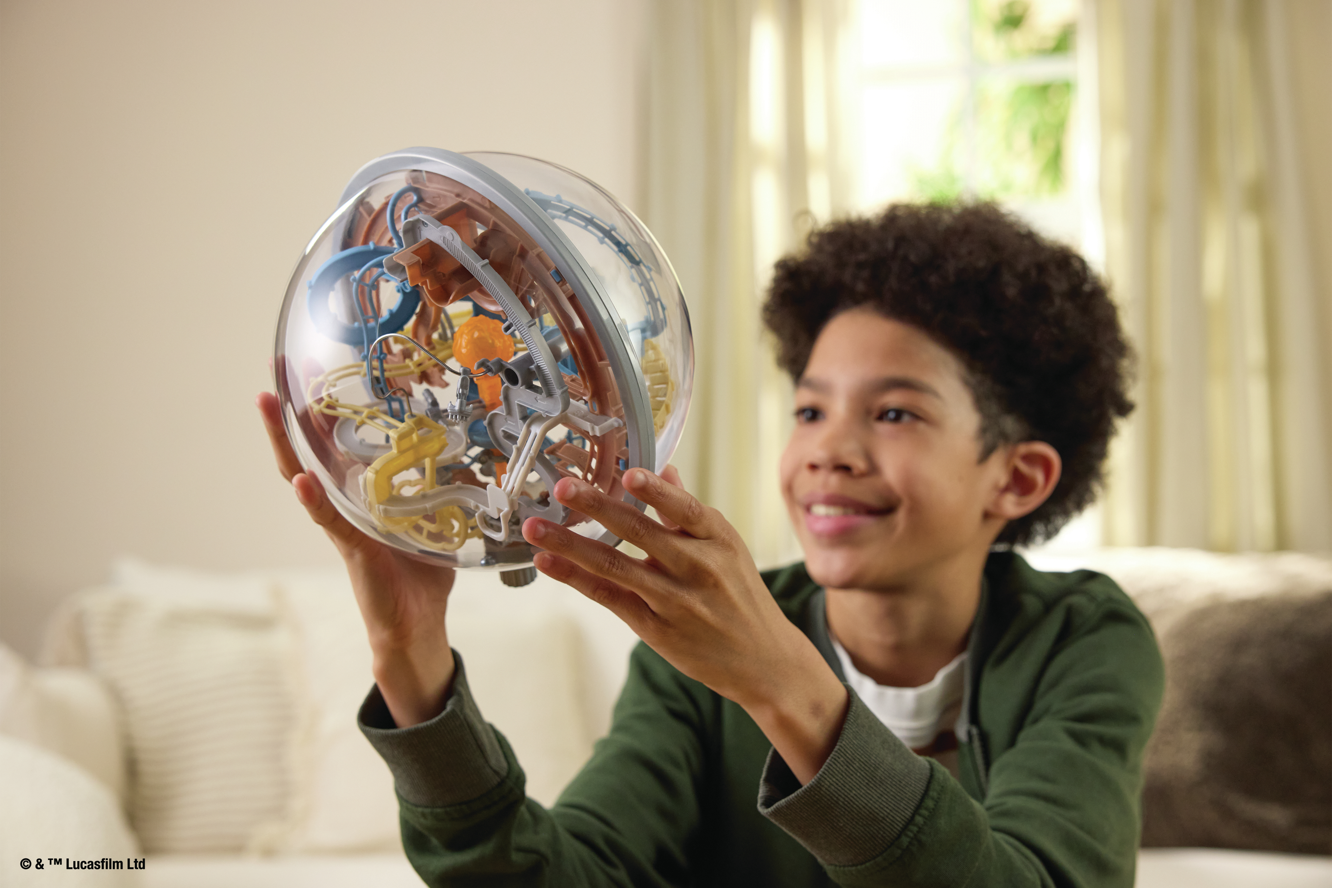 Chłopiec trzyma przezroczystą sferyczną zabawkę labirynt. Uśmiecha się, patrząc na zabawkę. Tło przedstawia pokój ze światłem.