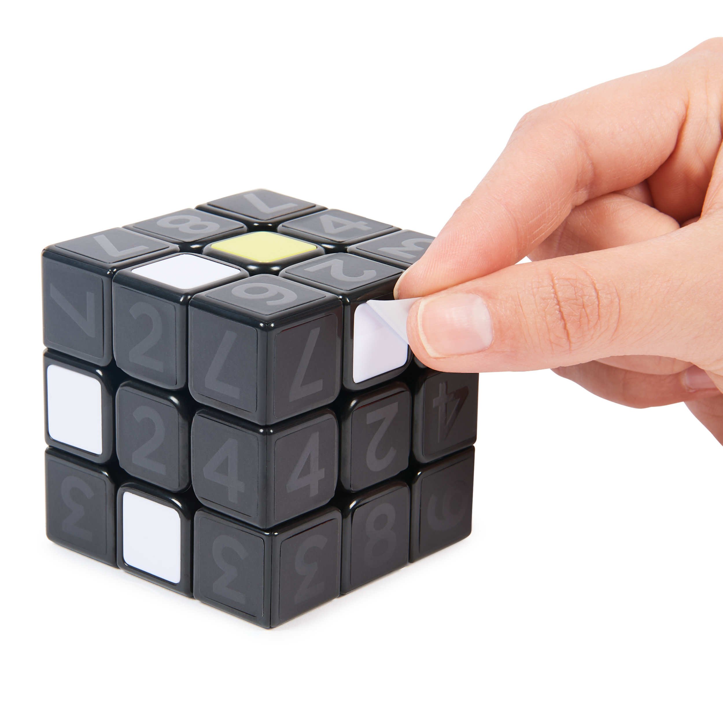 Una mano stacca un adesivo da un puzzle a cubo nero con numeri. Sfondo bianco. C'è un quadrato giallo in alto.