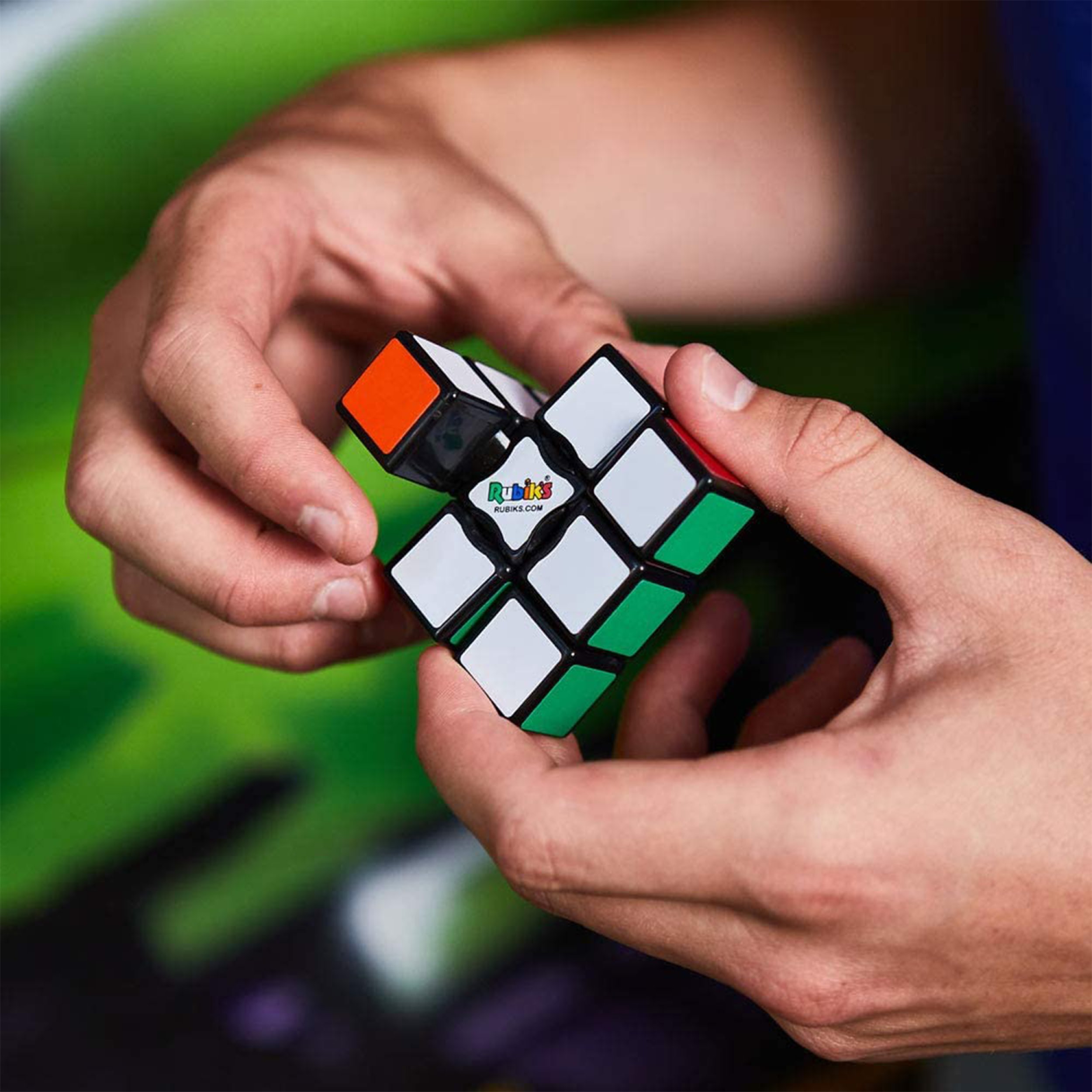 Hände halten einen Rubik's Cube, eine Seite orange. Andere Seiten zeigen weiße, grüne und rote Farben.