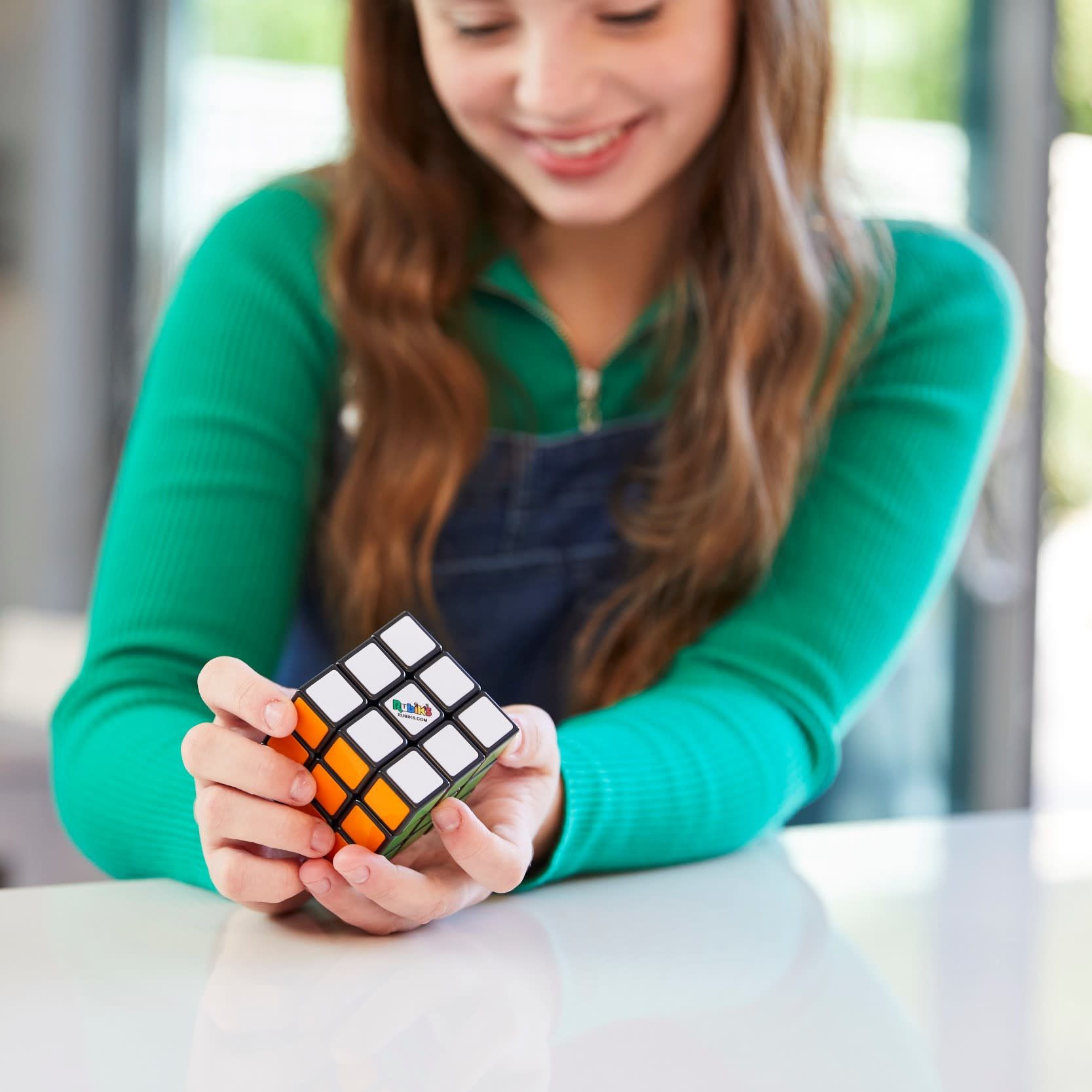 Ein lächelndes Mädchen hält einen Rubik's Cube. Der Würfel hat weiße, orange und grüne Farben. Das Mädchen trägt ein grünes Oberteil.