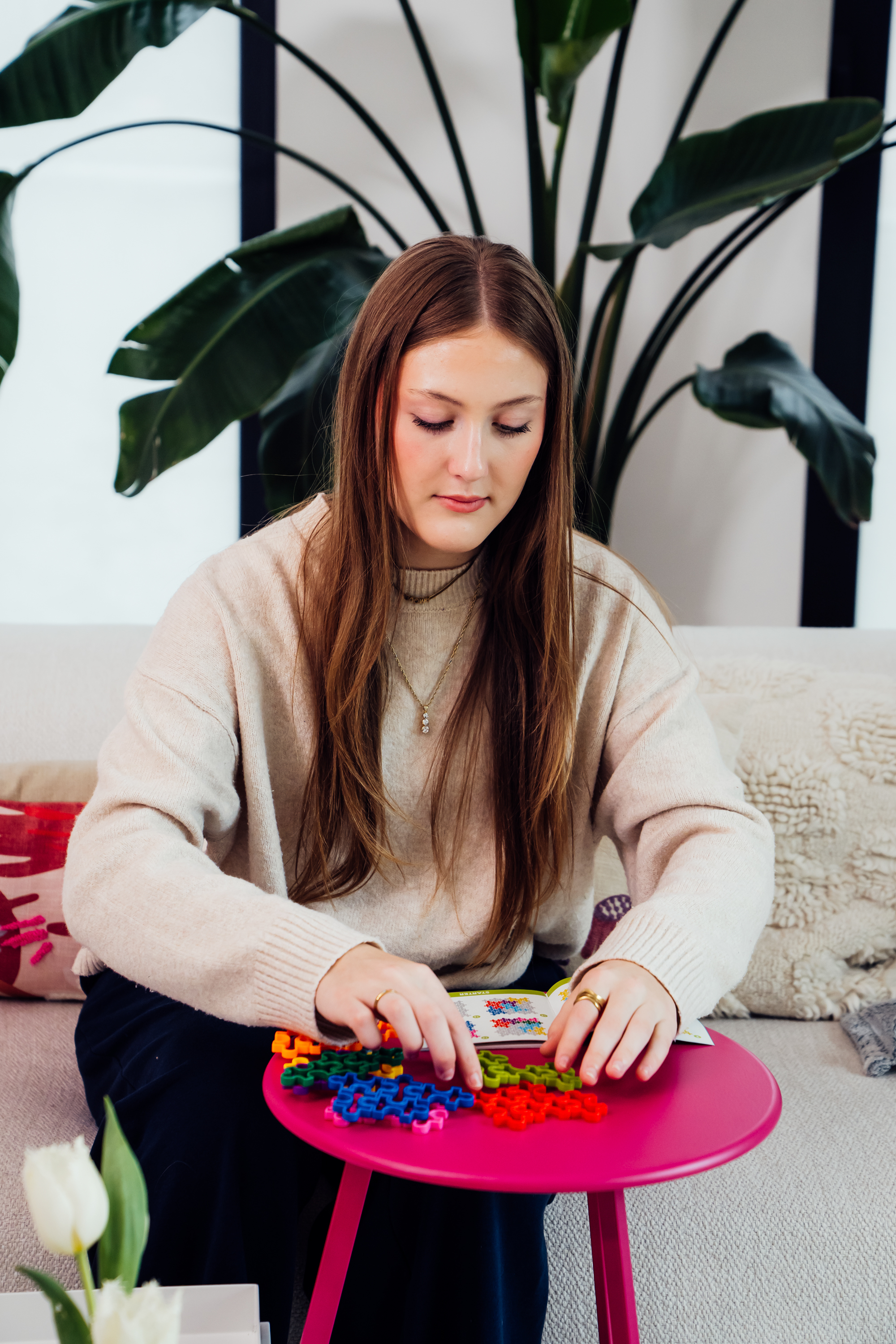 Kobieta siedzi, układając kolorowe elementy układanki na różowym stole. W tle roślina i kanapa.