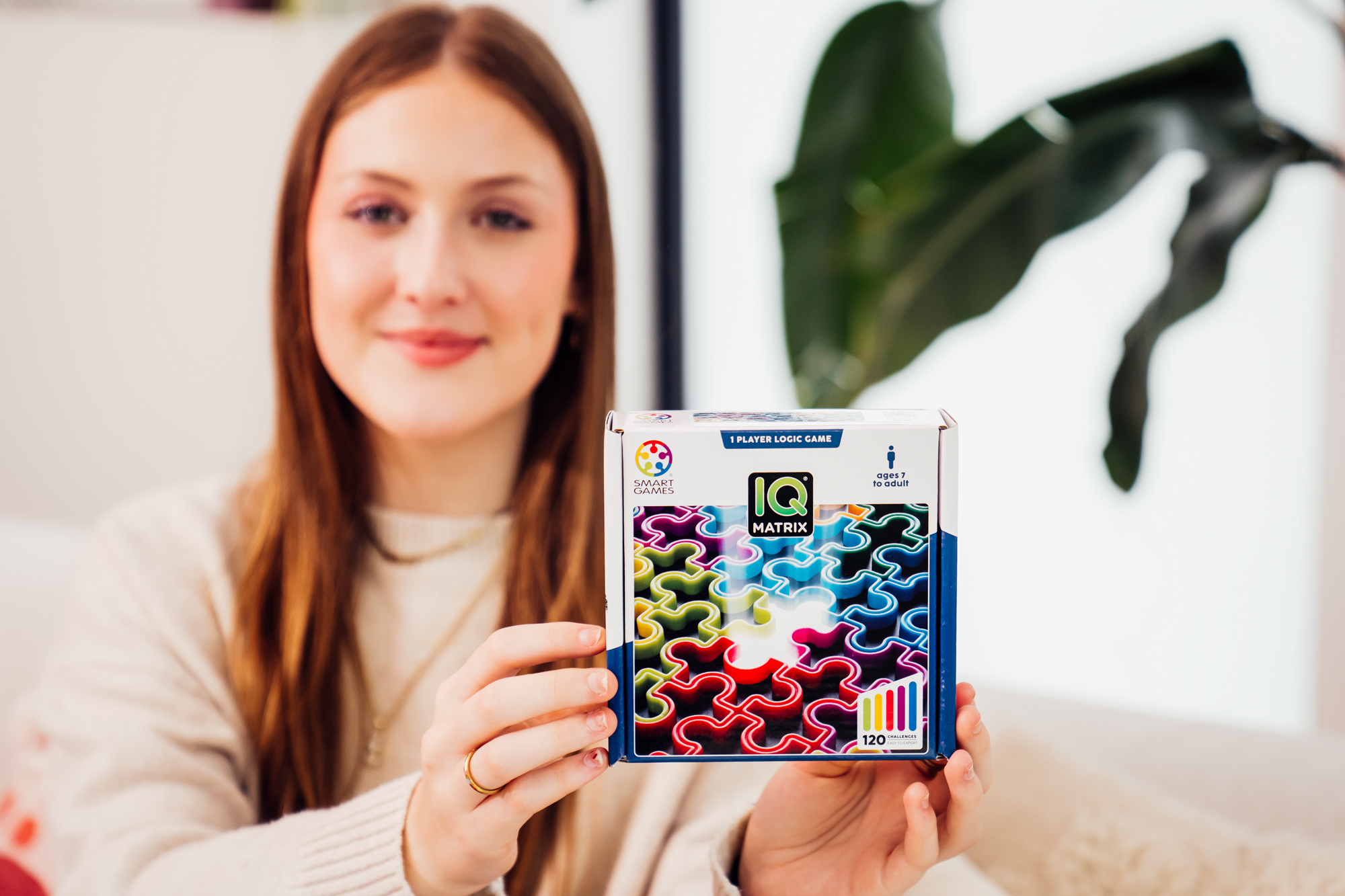 Kobieta trzymająca pudełko z puzzlami IQ Matrix. Pudełko ma kolorowe elementy układanki. Kobieta uśmiecha się, z pierścionkiem na palcu.