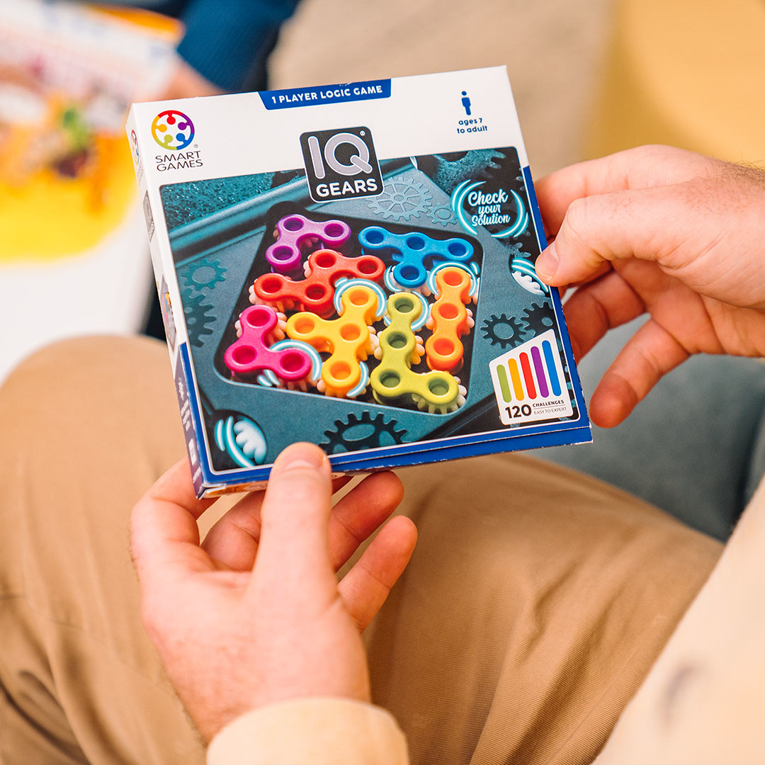 Eine Person hält eine IQ Gears Puzzlebox. Die Box zeigt bunte Zahnräder und das Smart Games Logo. Hände und Kleidung sind sichtbar.