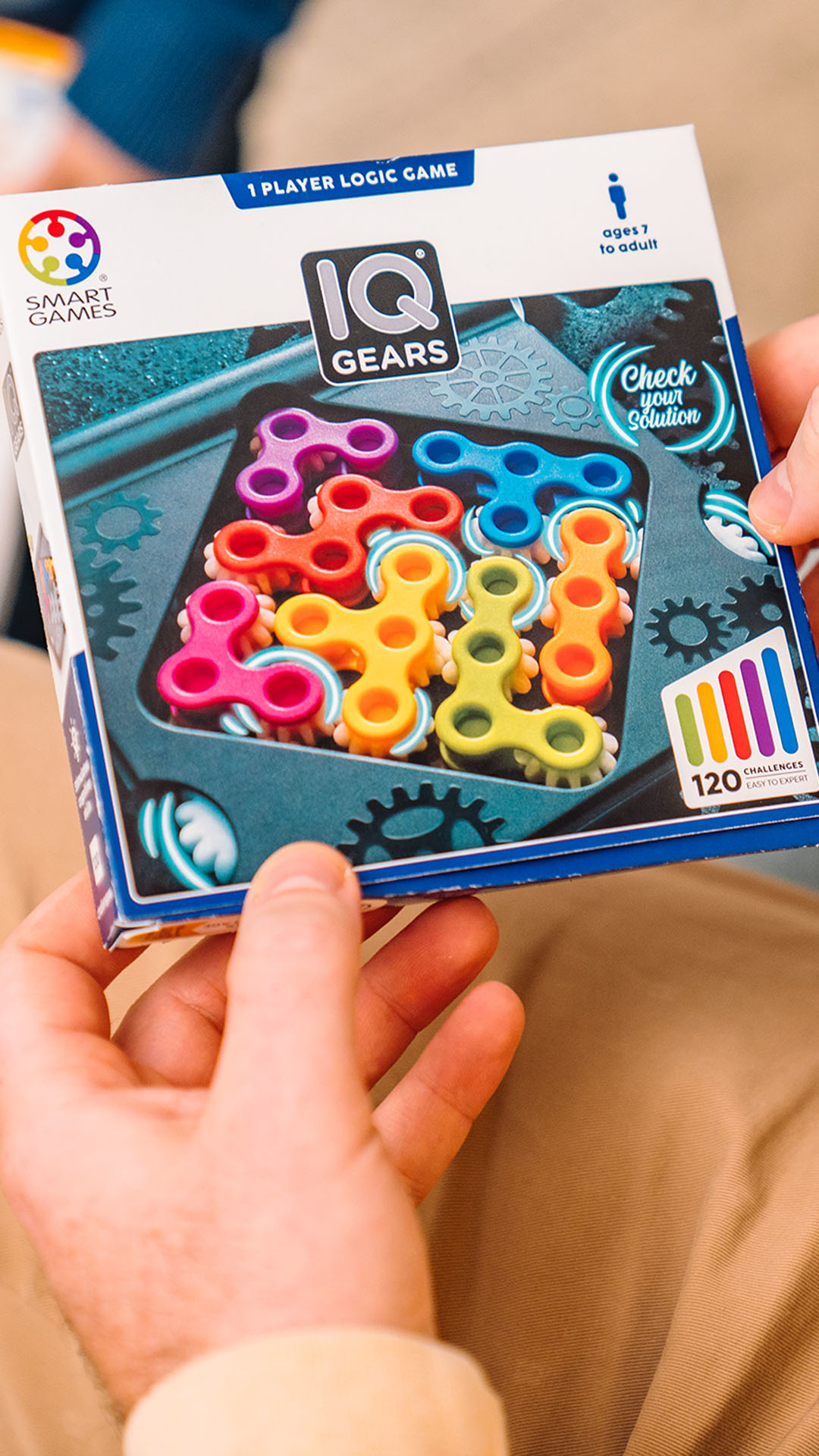 Eine Person hält eine IQ Gears-Spielbox. Die Box hat bunte Zahnräder im Inneren. Der Hintergrund ist die Kleidung einer Person.
