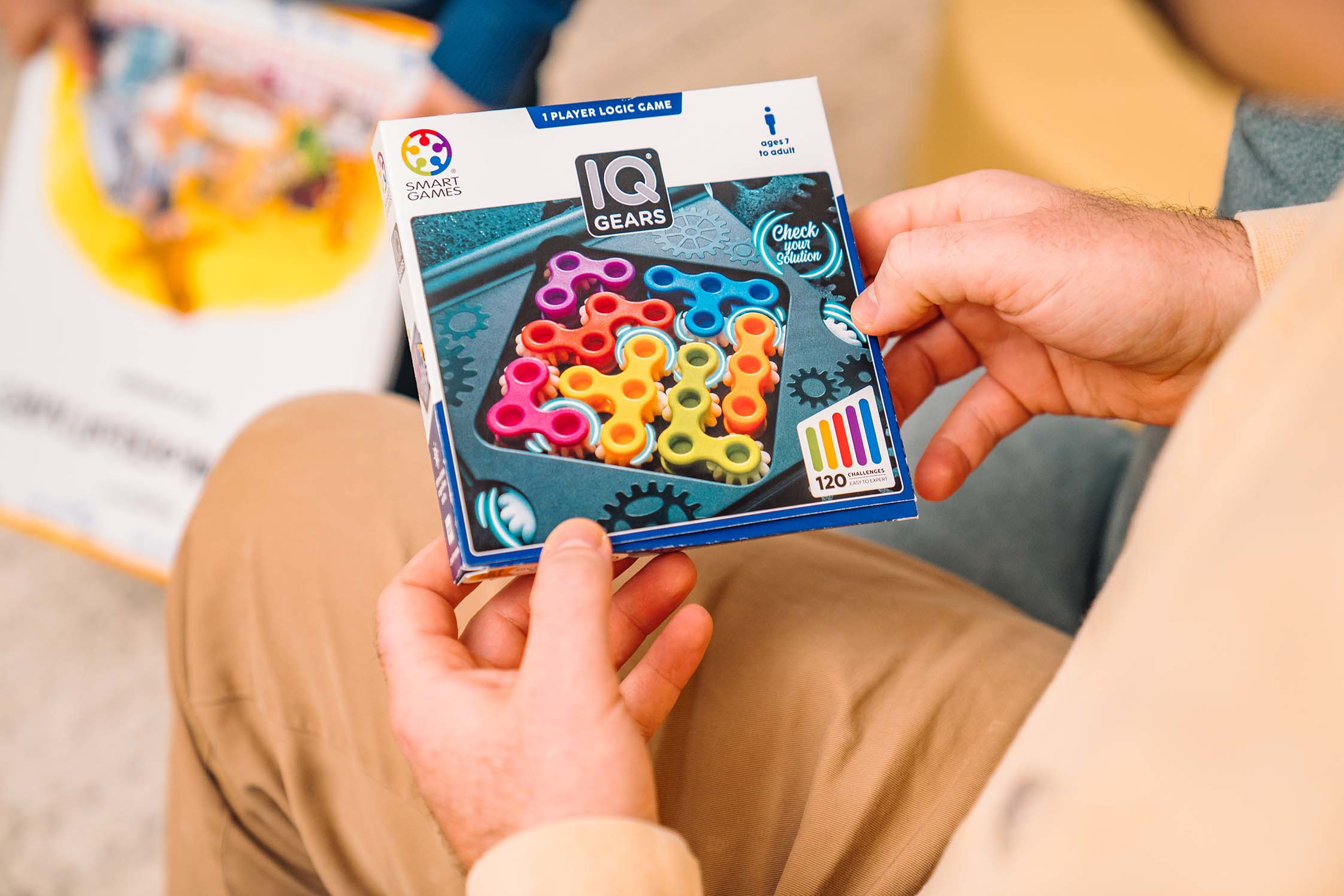 Eine Person hält eine IQ Gears Puzzlebox. Die Box zeigt farbige Zahnräder und sagt ab 7 Jahren. Andere Person im Hintergrund.