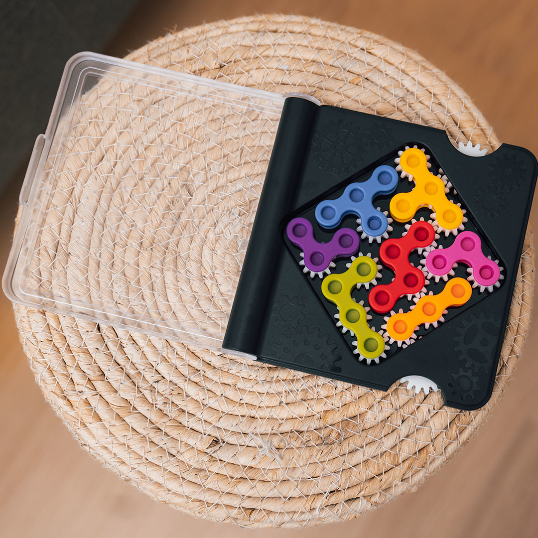 Ein farbenfrohes Puzzle mit ineinandergreifenden Zahnrädern liegt auf einer gewebten Oberfläche. Ein klarer Plastikdeckel ist daneben geöffnet.
