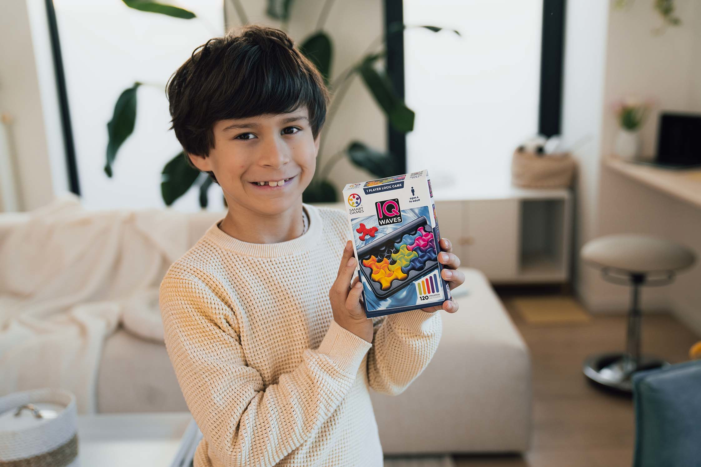 Ein lächelnder Junge hält ein Puzzlespiel. Die Spielbox ist sichtbar. Im Hintergrund sind ein Sofa und eine Pflanze zu sehen.
