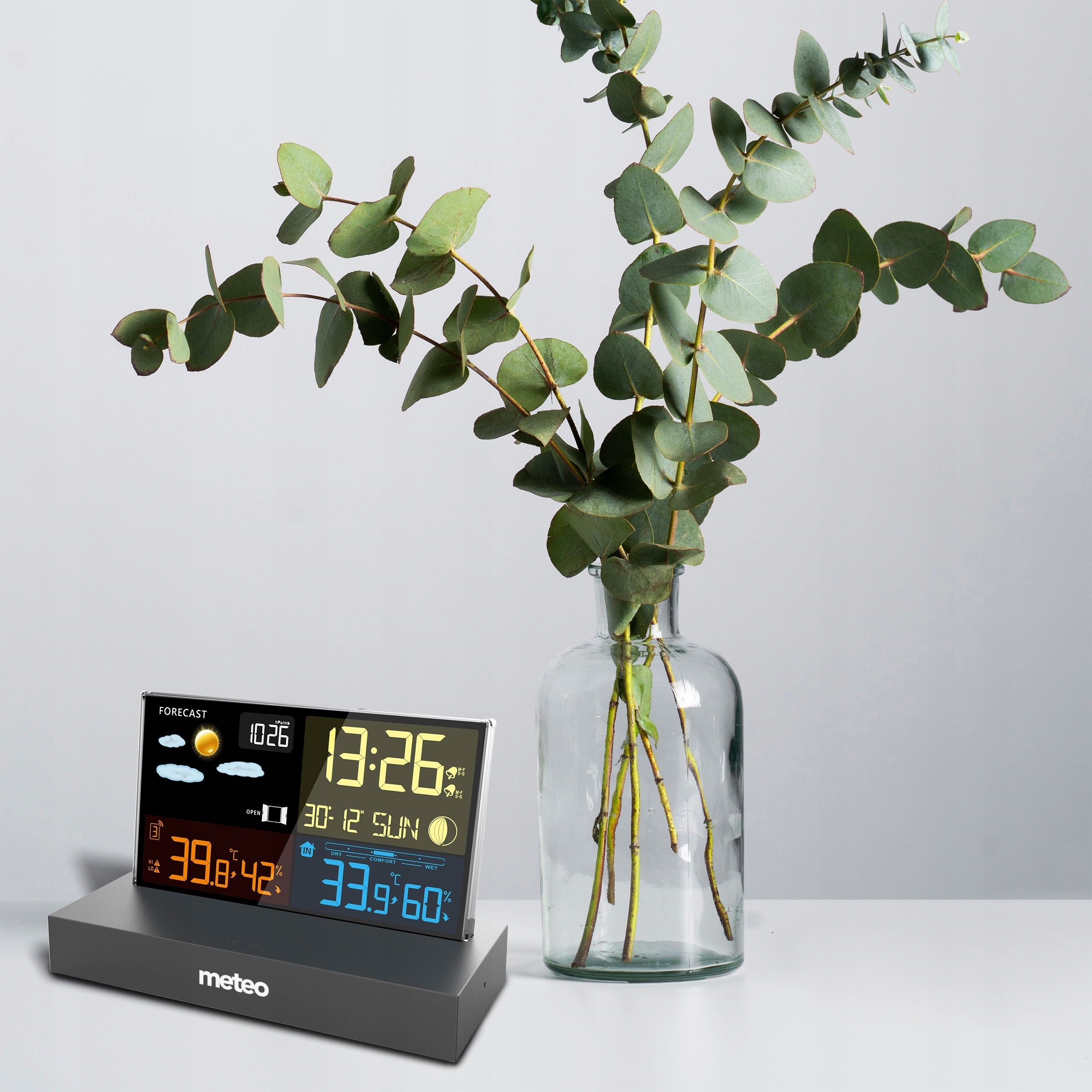 Stacja pogodowa wyświetla godzinę i temperaturę. Wazon z zielonymi liśćmi stoi na białej powierzchni, na szarym tle.