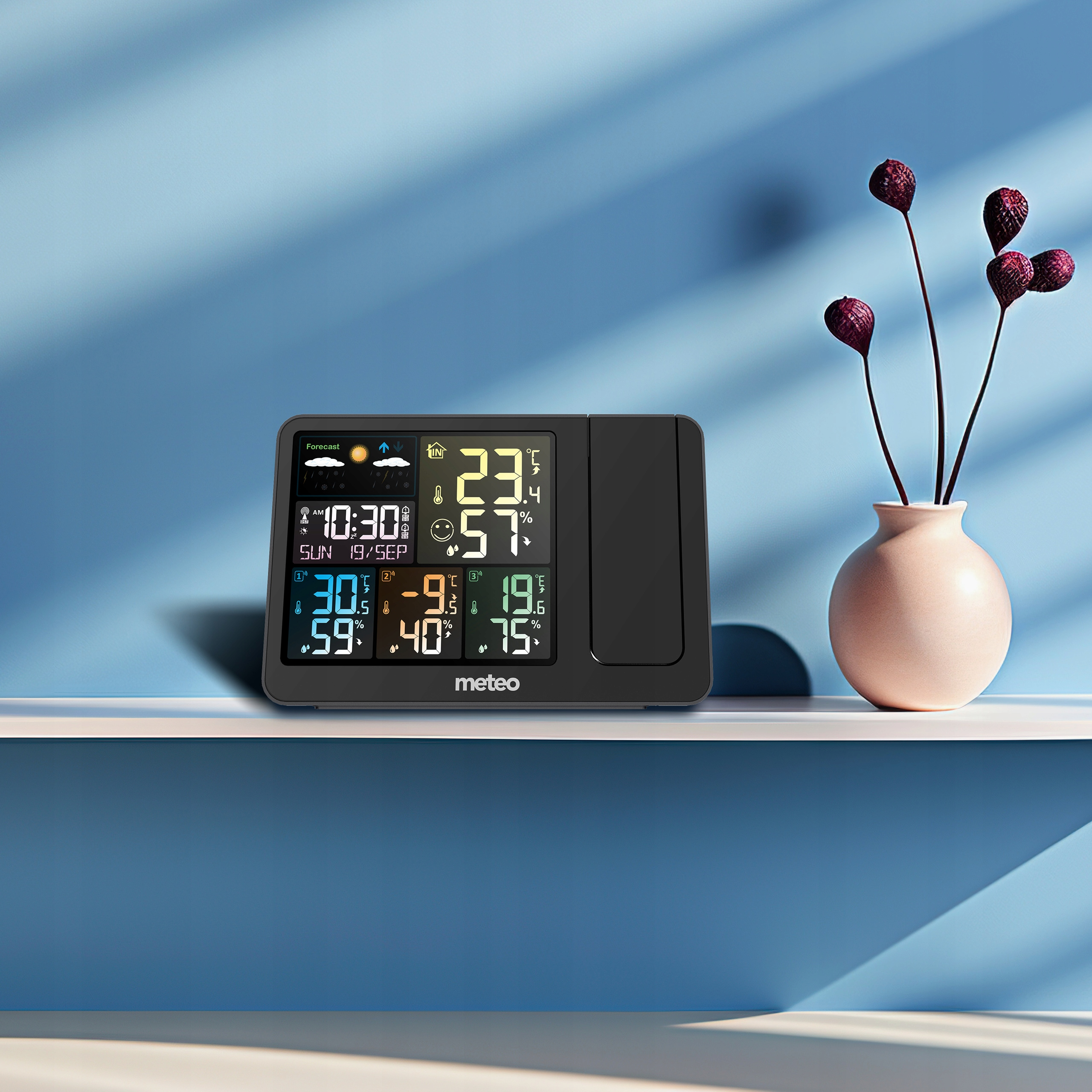 Stacja pogodowa wyświetla temperaturę i wilgotność. Po prawej wazon z czerwonymi kwiatami.