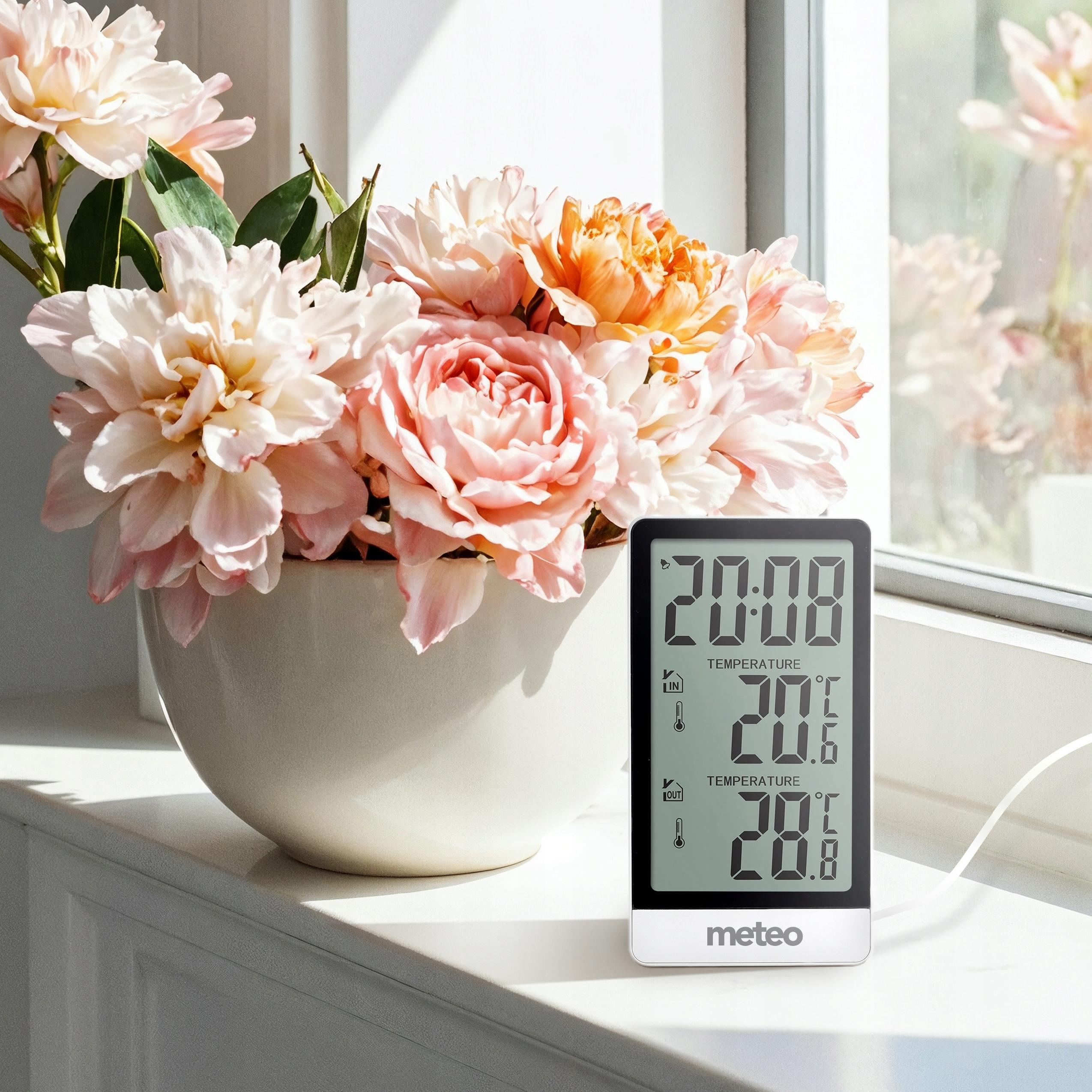 Wazon z różowymi i pomarańczowymi kwiatami stoi obok cyfrowego termometru na parapecie.