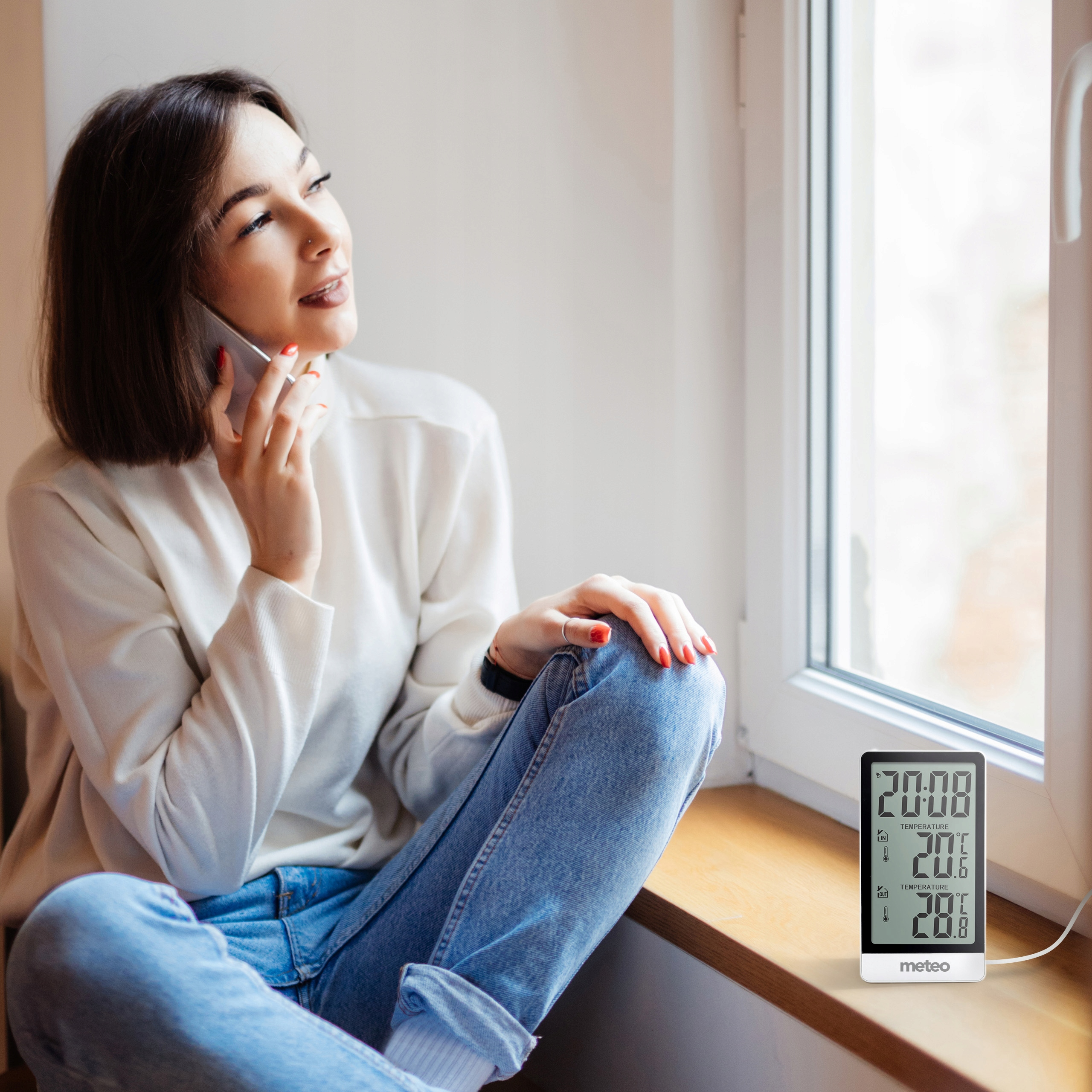 Kobieta przez telefon przy oknie, w dżinsach i swetrze. Termometr na parapecie.