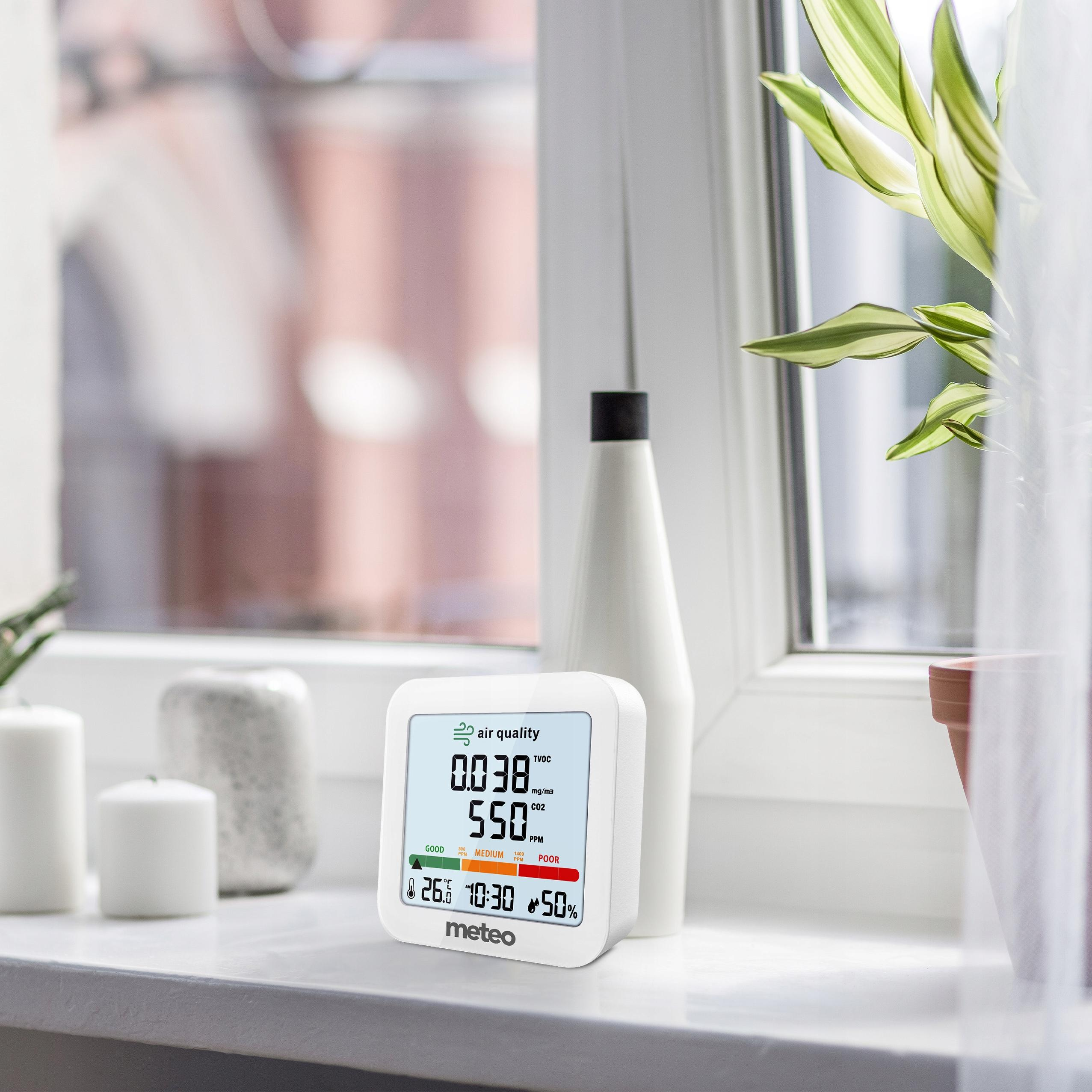 Monitor jakości powietrza pokazuje odczyty. W tle okno z roślinami i świecami.