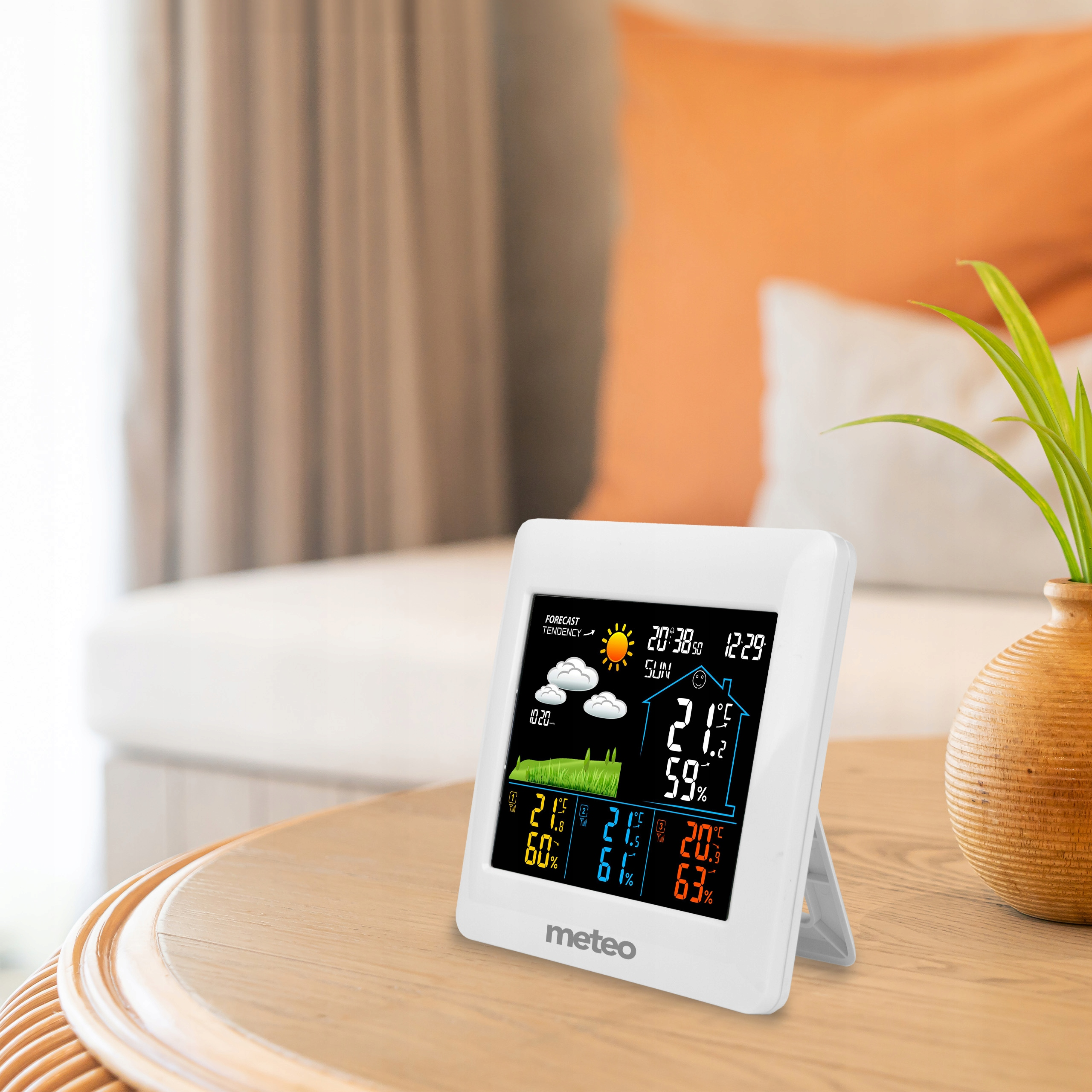 Biała stacja pogodowa wyświetla czas, temperaturę i prognozę. Stoi na drewnianym stole, w tle pomarańczowa poduszka.