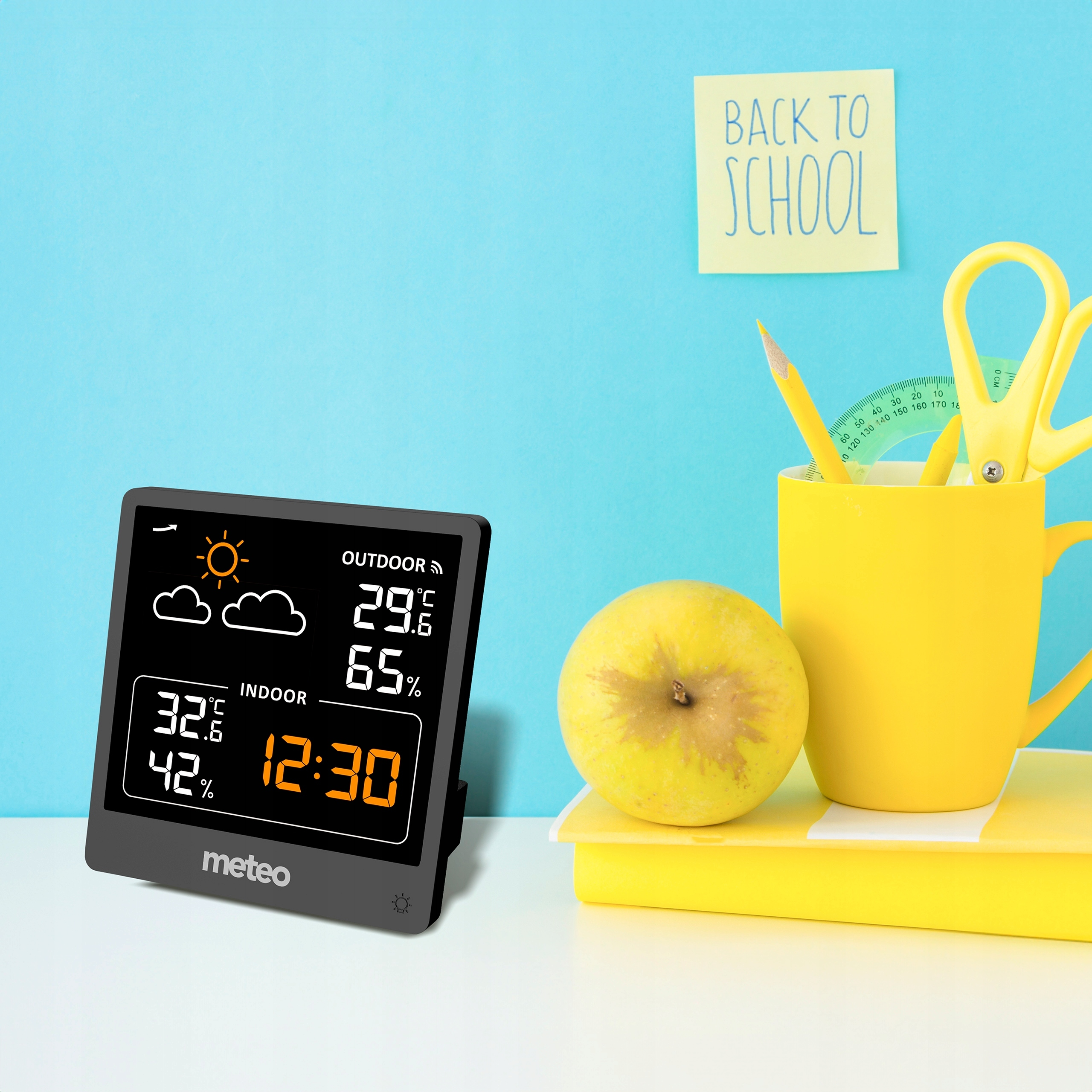 Stacja pogodowa wyświetla temperatury zewnętrzne/wewnętrzne. Żółty kubek zawiera przybory szkolne. Jabłko i notatka 'powrót do szkoły'.