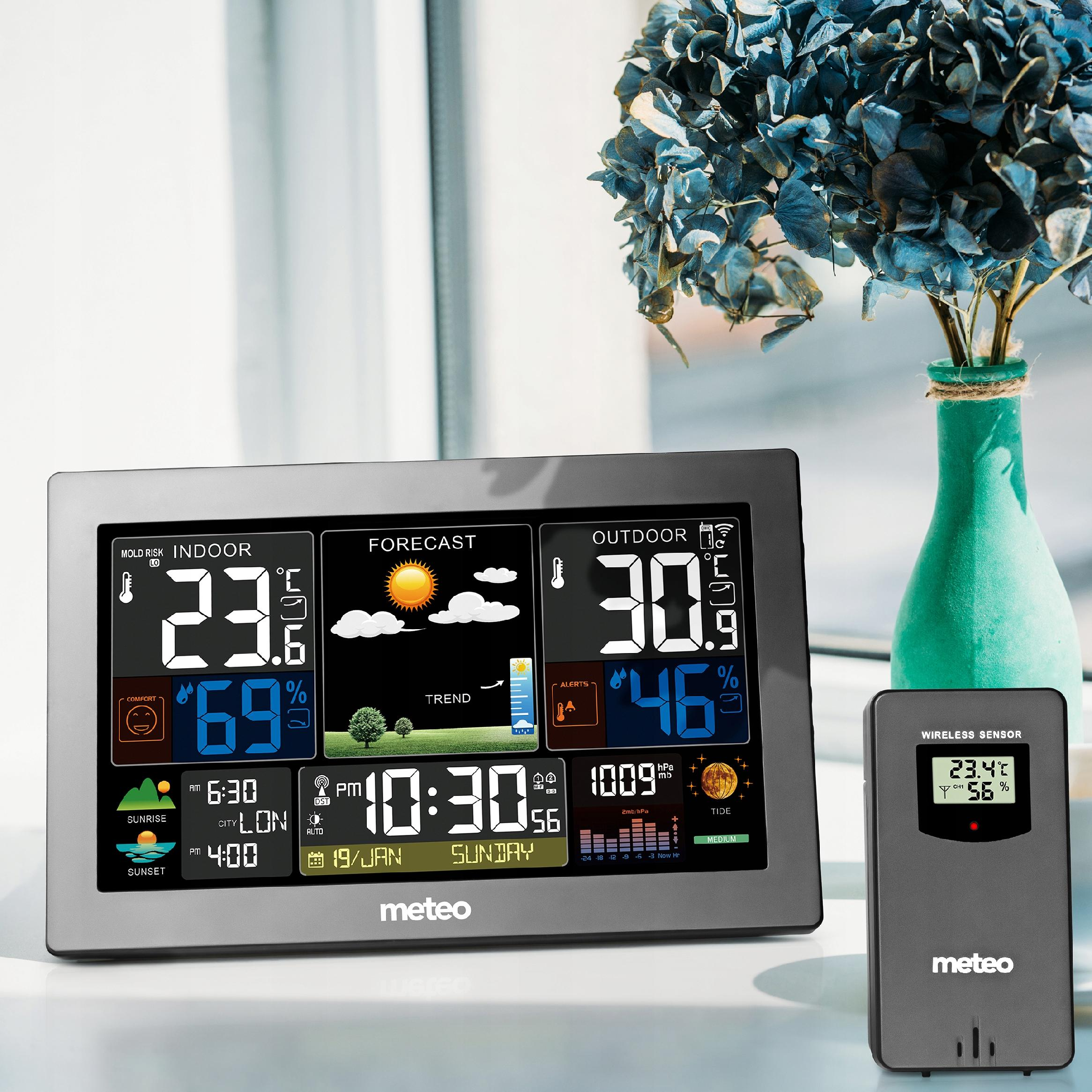 Stacja pogodowa wyświetla temperaturę wewnątrz/na zewnątrz, prognozę, czas i wilgotność. W wazonie są niebieskie kwiaty.