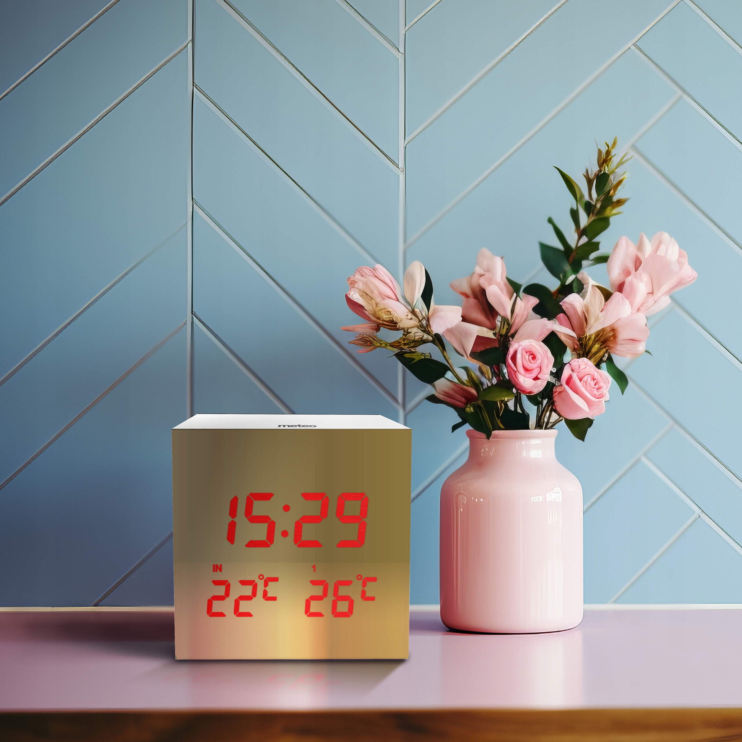 Złoty zegar wyświetlający godzinę i temperaturę stoi na różowej powierzchni z wazonem różowych kwiatów i niebieską ścianą.
