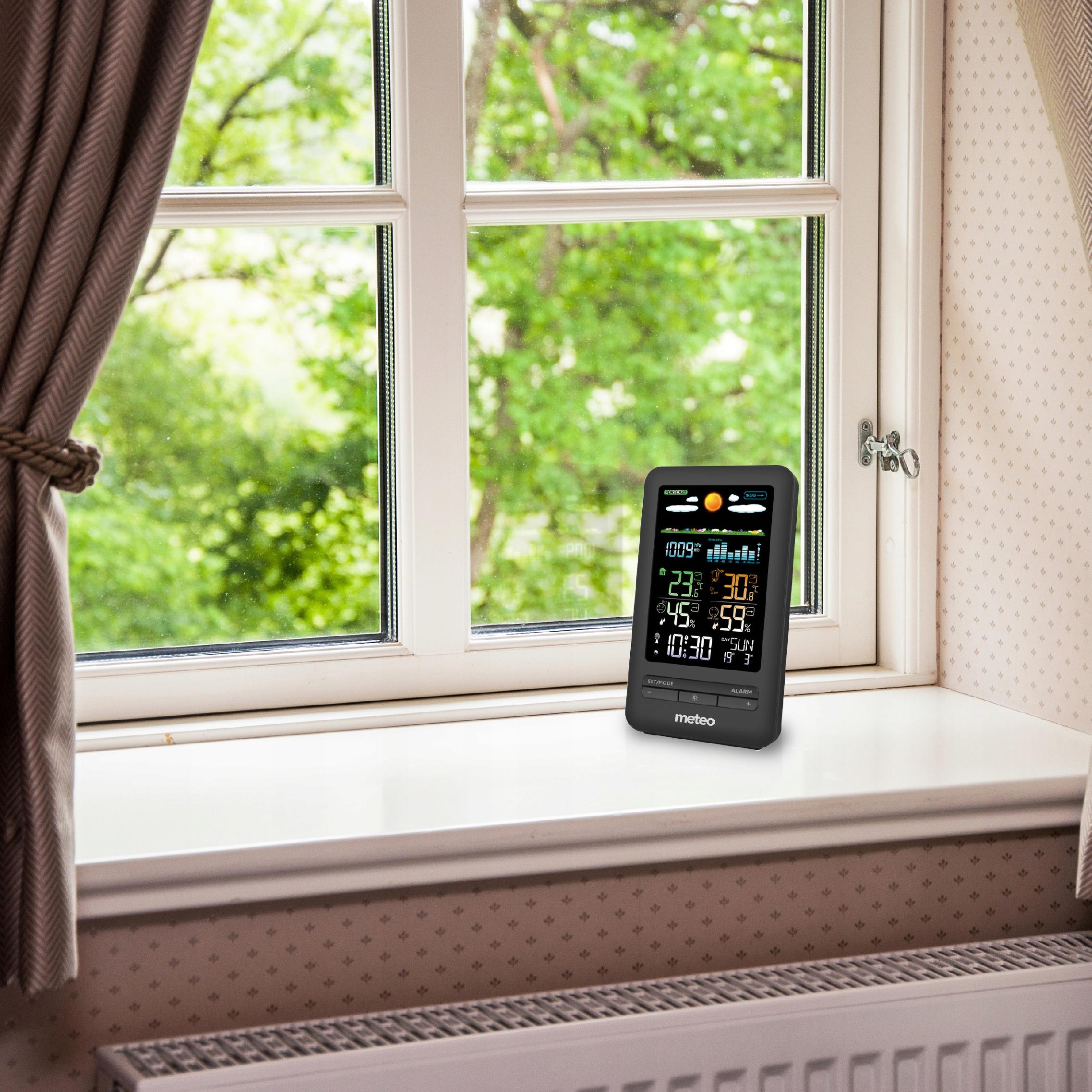 Stacja pogodowa na parapecie. Okno z drzewami w tle. Zasłony po lewej.