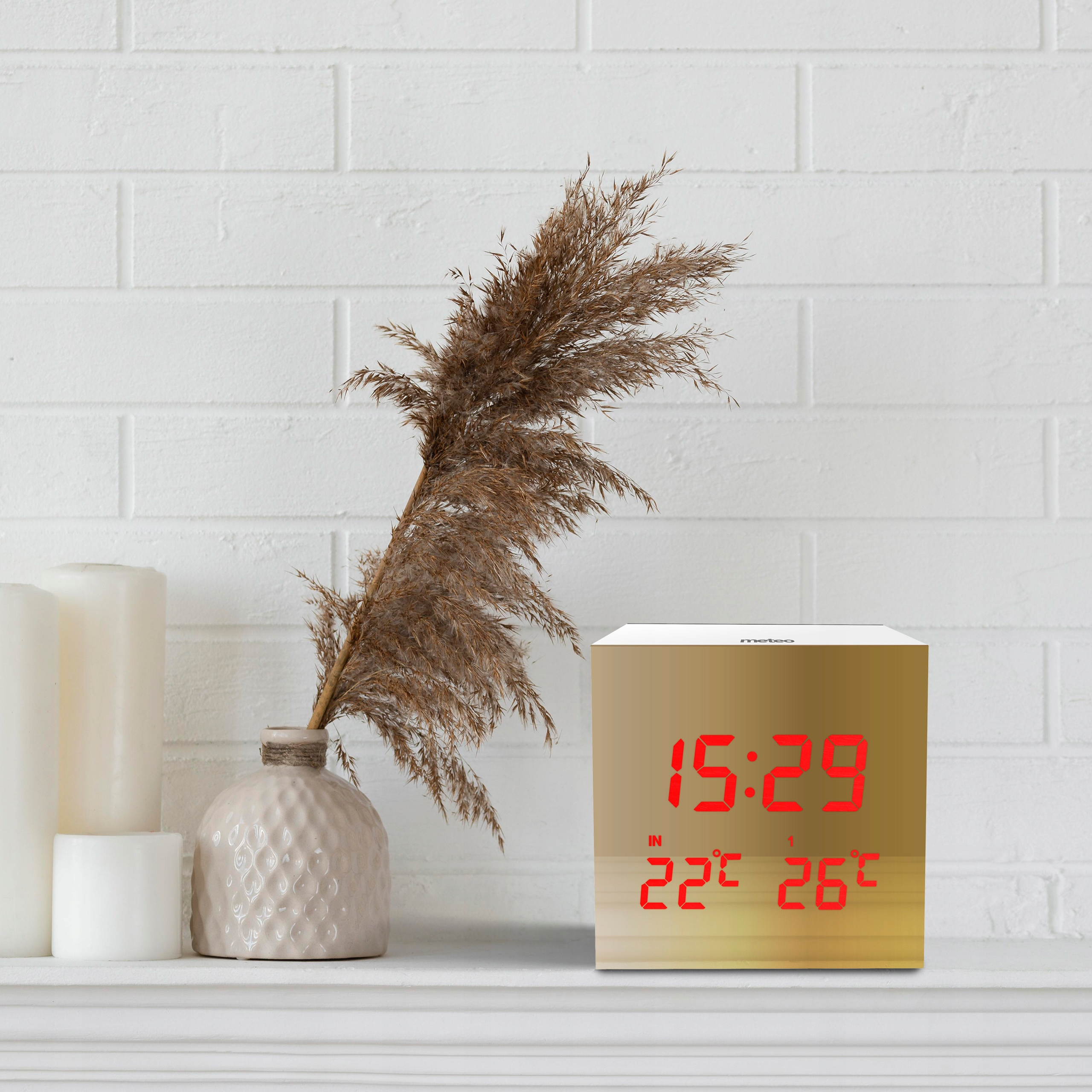Zegar pokazujący 15:29 i temperatury. Obok wazon z suchymi roślinami, świecami i ceglanym murem.