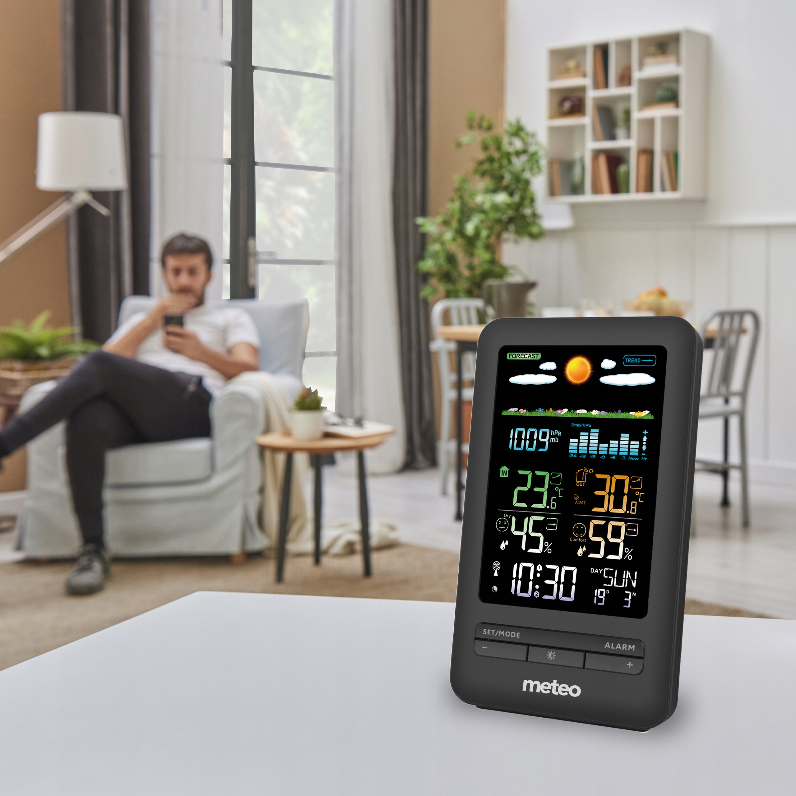 Stacja pogodowa wyświetla dane na stole. Mężczyzna siedzi na sofie w rozmytym tle. Po lewej stronie jest okno.