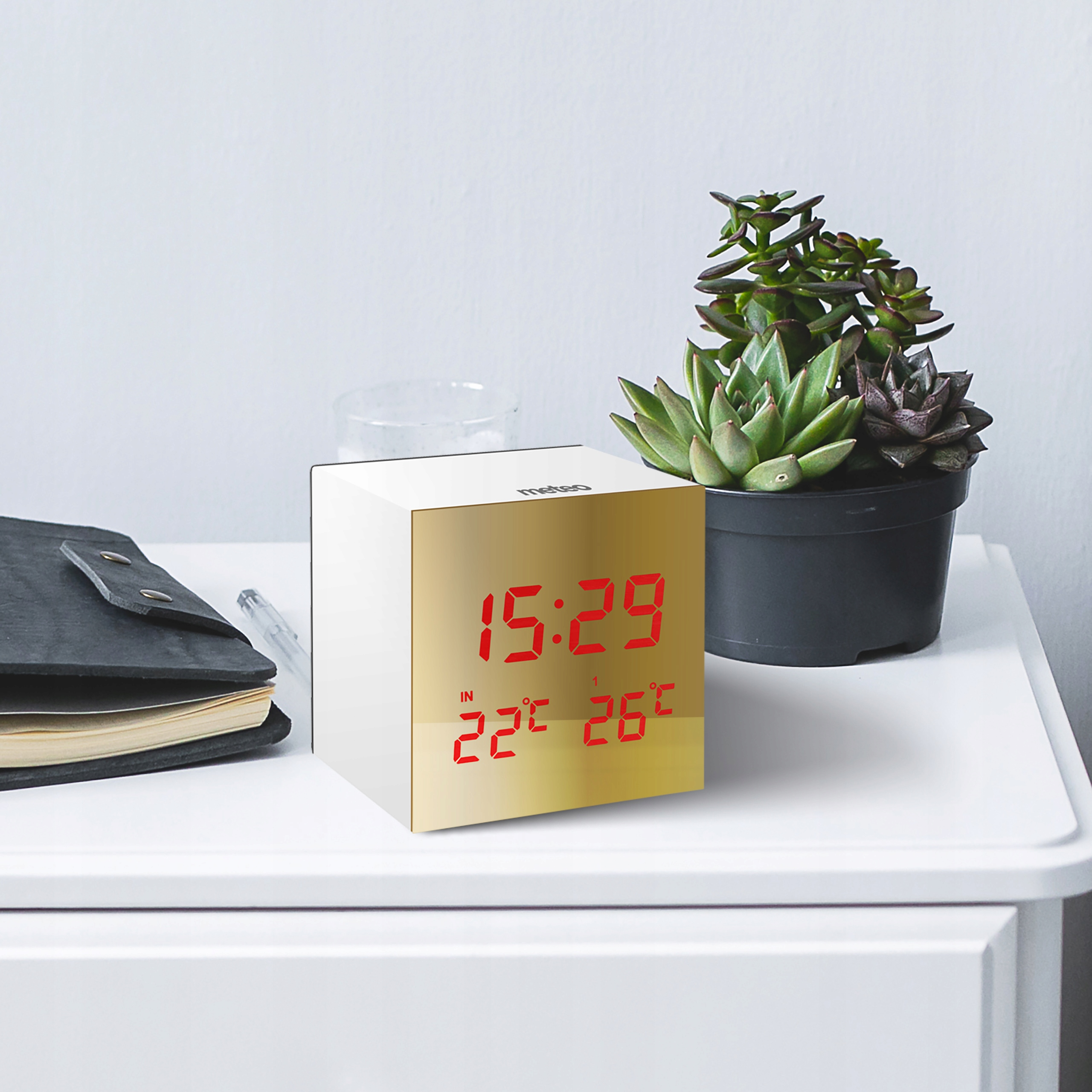 Biała szafka nocna z zegarem, rośliną, notatnikiem i długopisem. Zegar wyświetla godzinę i temperaturę na czerwono.