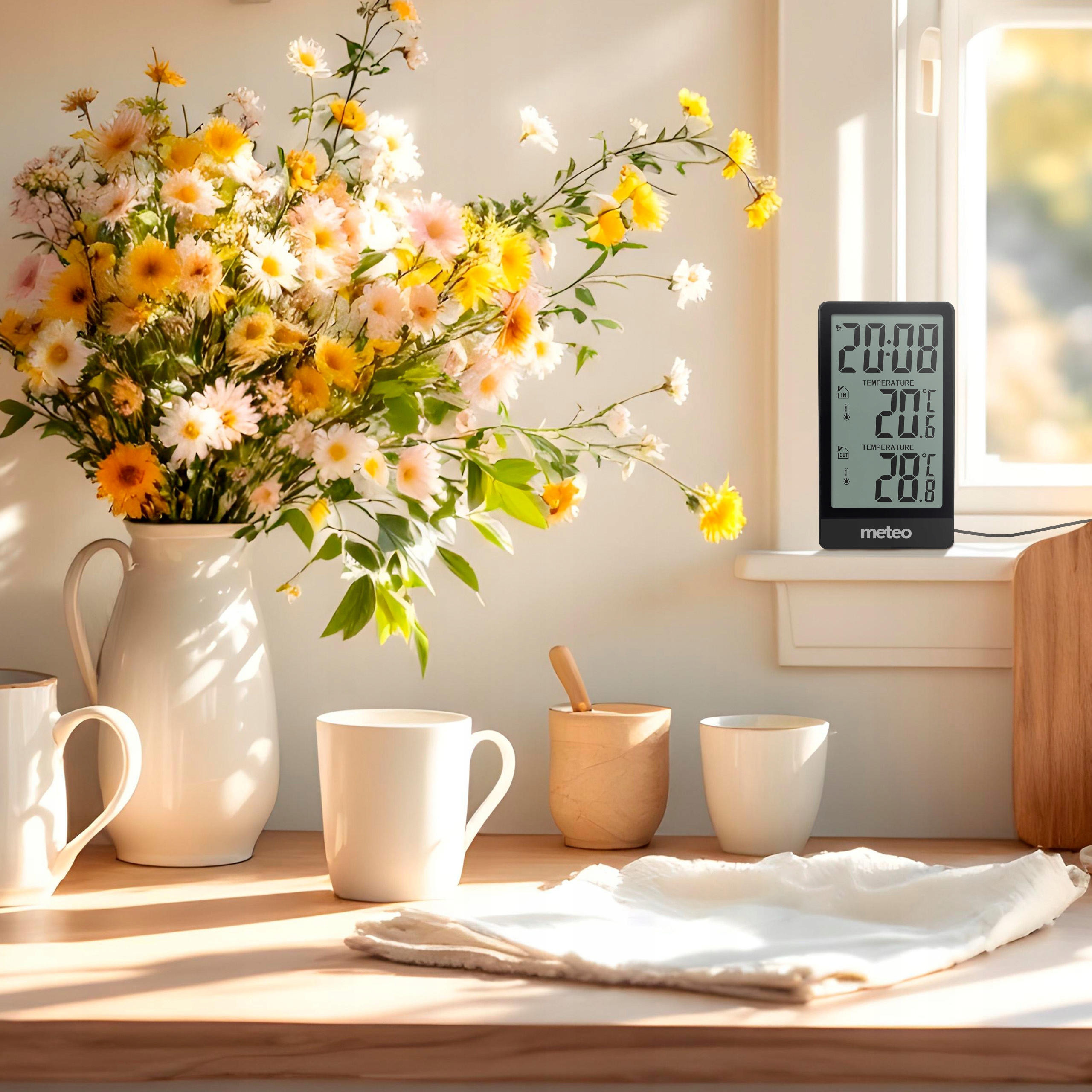 Scena kuchenna z kwiatami w wazonie, filiżankami i termometrem. Światło słoneczne wpada przez okno.