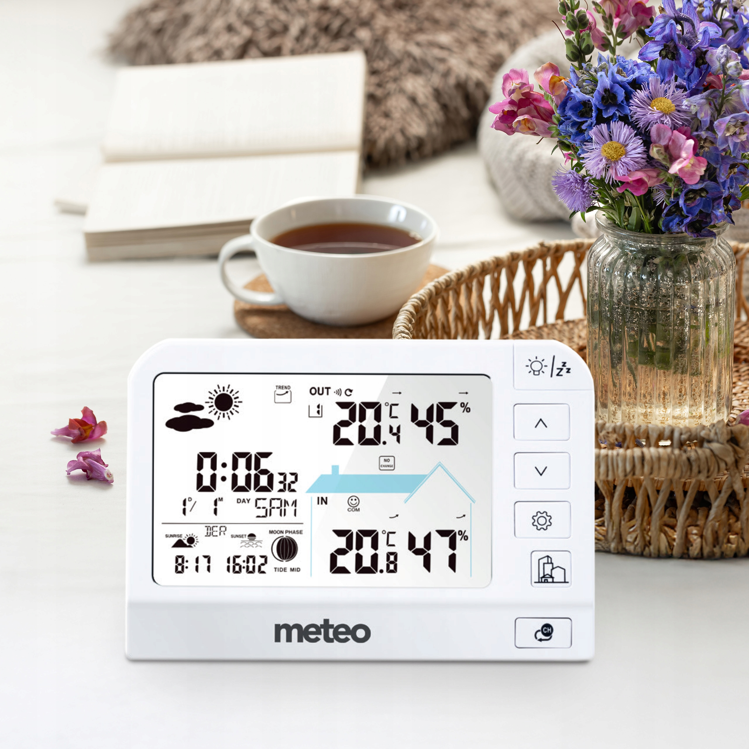 Biała stacja pogodowa wyświetla temperaturę i godzinę. Kwiaty w wazonie, filiżanka herbaty i książka na stole.