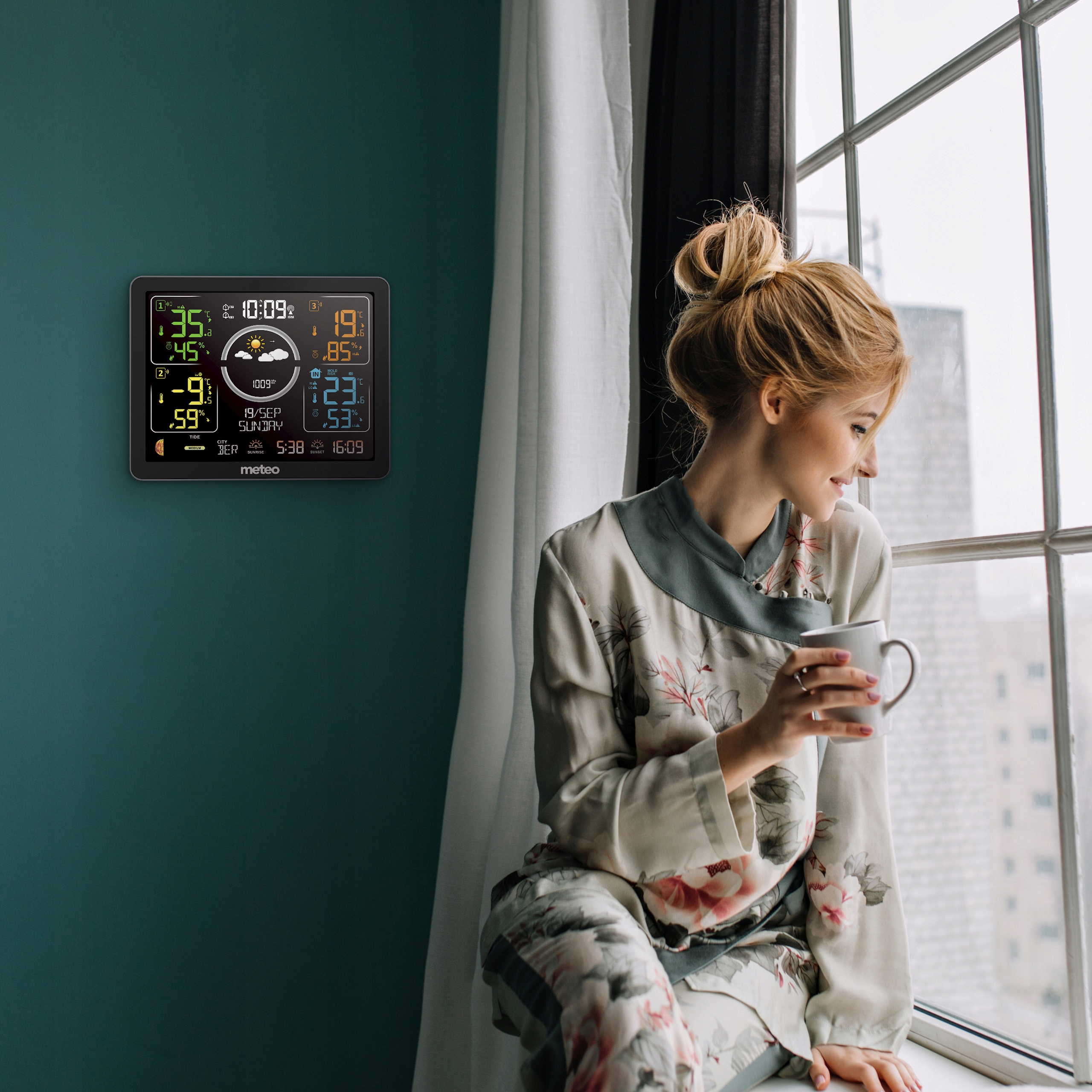 Kobieta z kubkiem patrzy przez okno. Stacja pogodowa na ścianie pokazuje temperaturę i godzinę. Widoczne okno i zasłony.