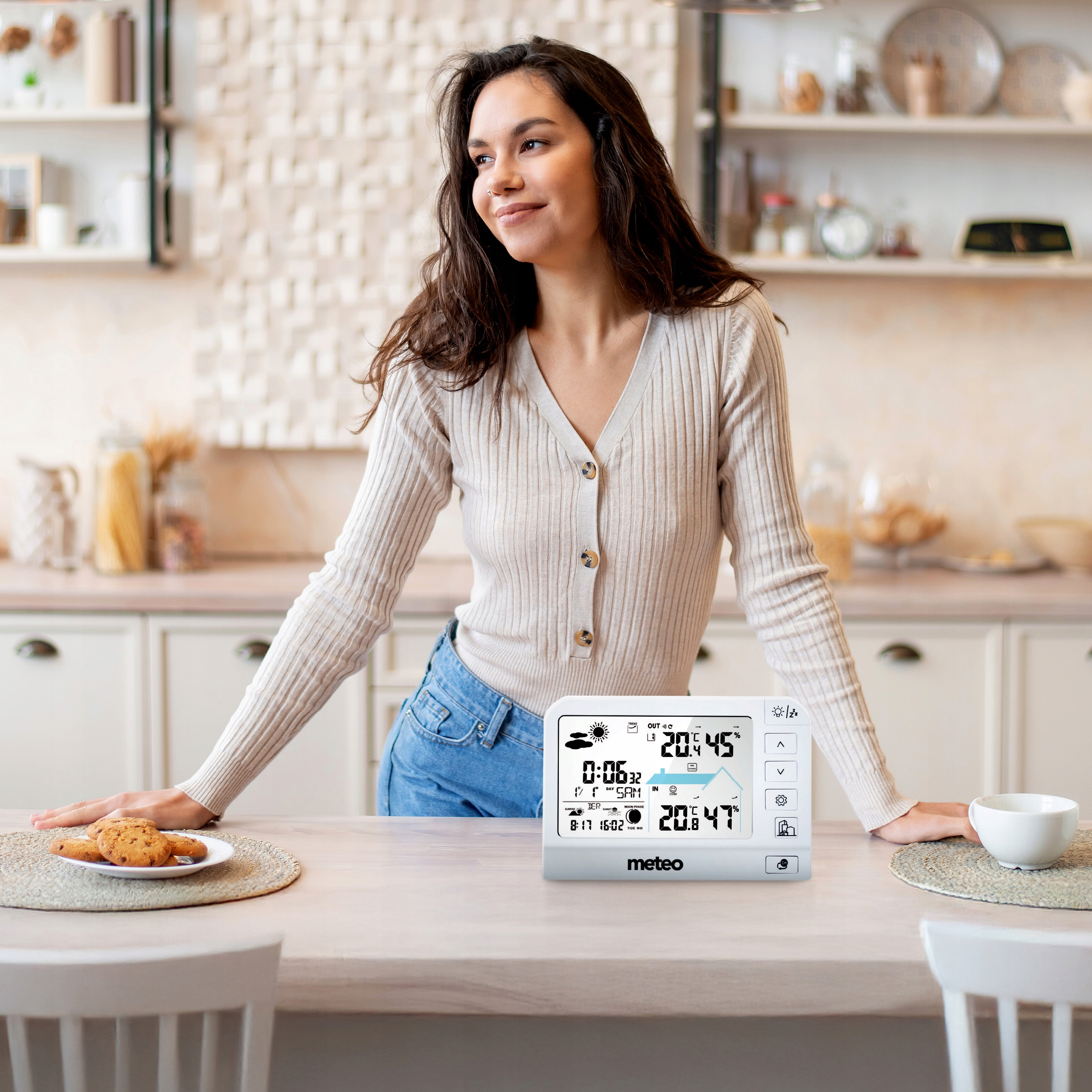 Kobieta w kuchni ze stacją pogodową. Ciastka i filiżanka na stole. Kobieta patrzy w bok uśmiechając się.