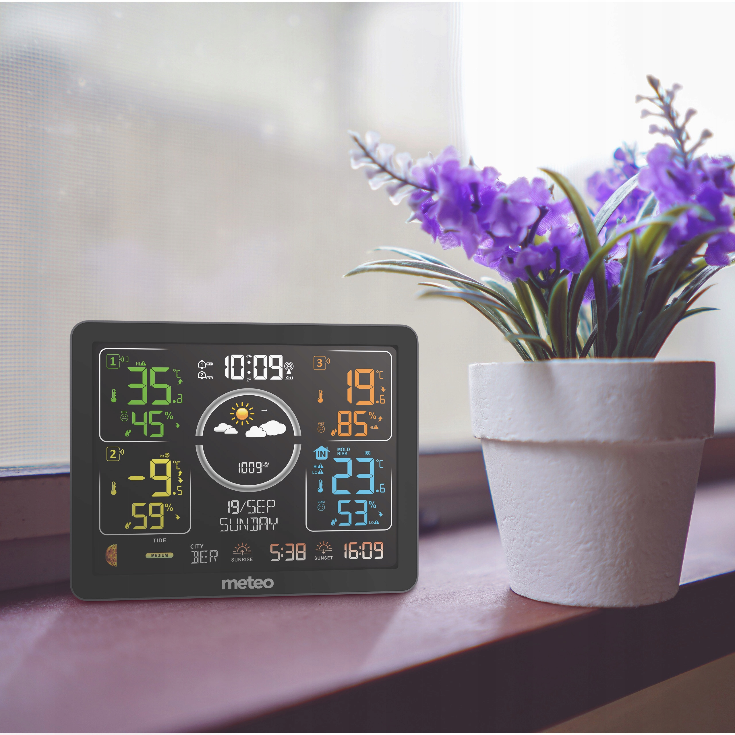 Stacja pogodowa wyświetla temperaturę, wilgotność i datę. Wazon z fioletowymi kwiatami na drewnianej powierzchni.