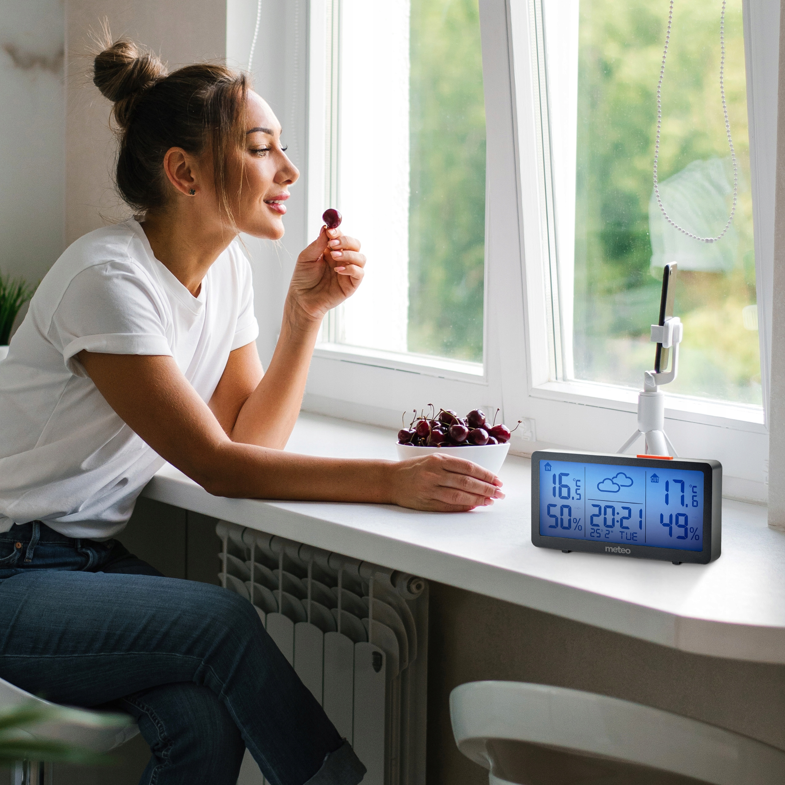 Kobieta jedząca wiśnię przy oknie. Wiśnie w misce. Stacja pogodowa na parapecie. Uchwyt na telefon.