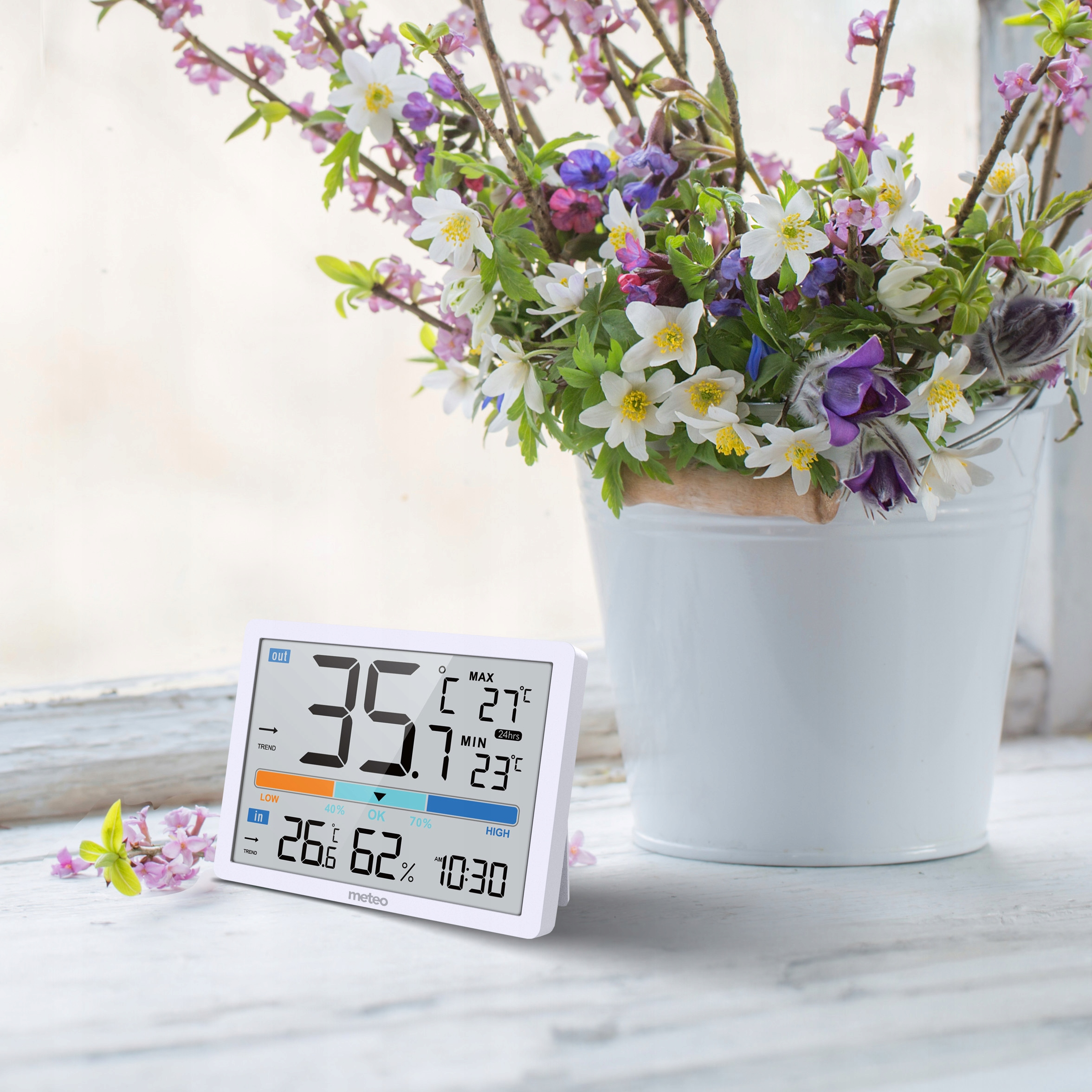 Biała cyfrowa stacja pogodowa wyświetla temperaturę i wilgotność. Bukiet kwiatów jest w białym wiadrze.