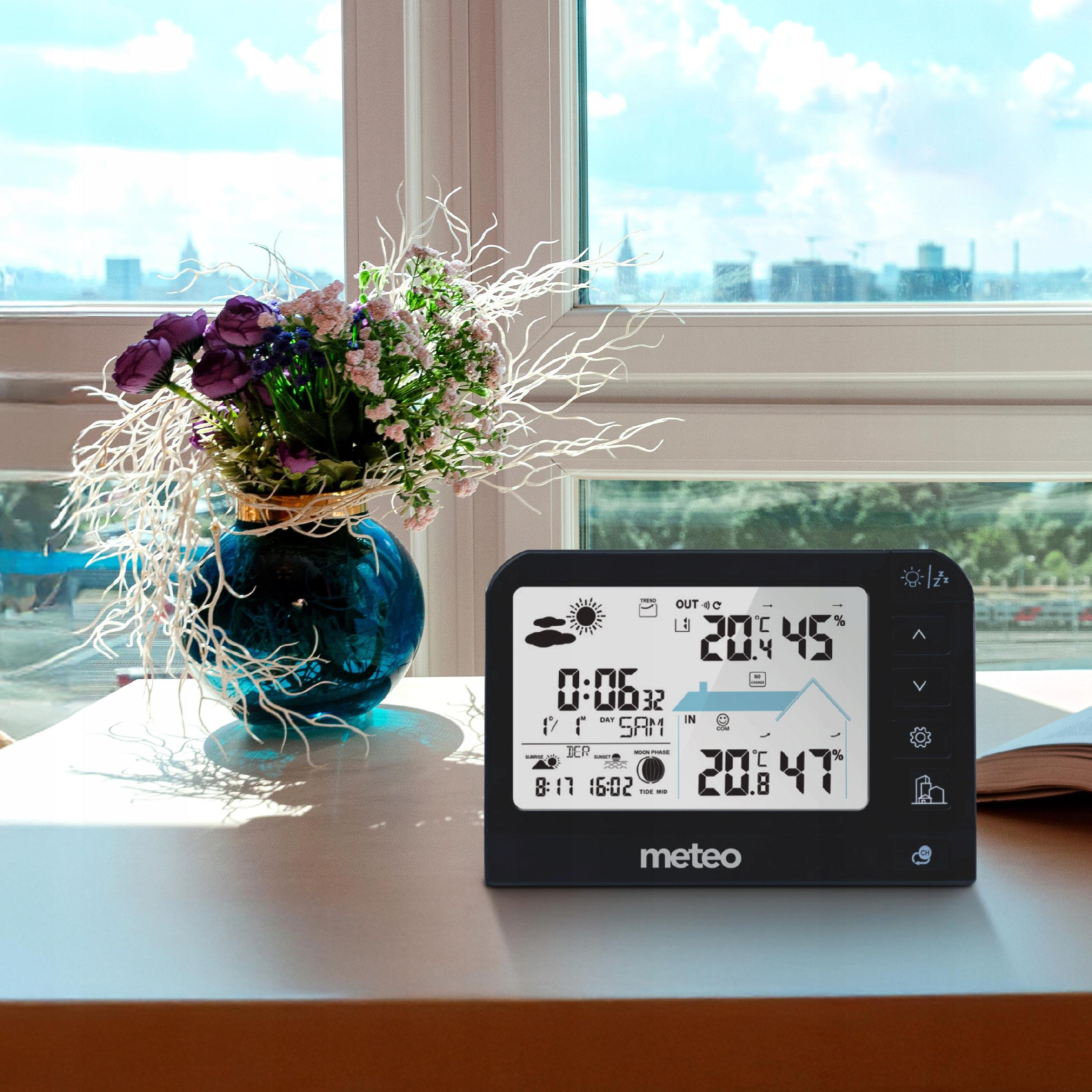 Stół z wazonem kwiatów, stacją pogodową i książką w pobliżu okna.