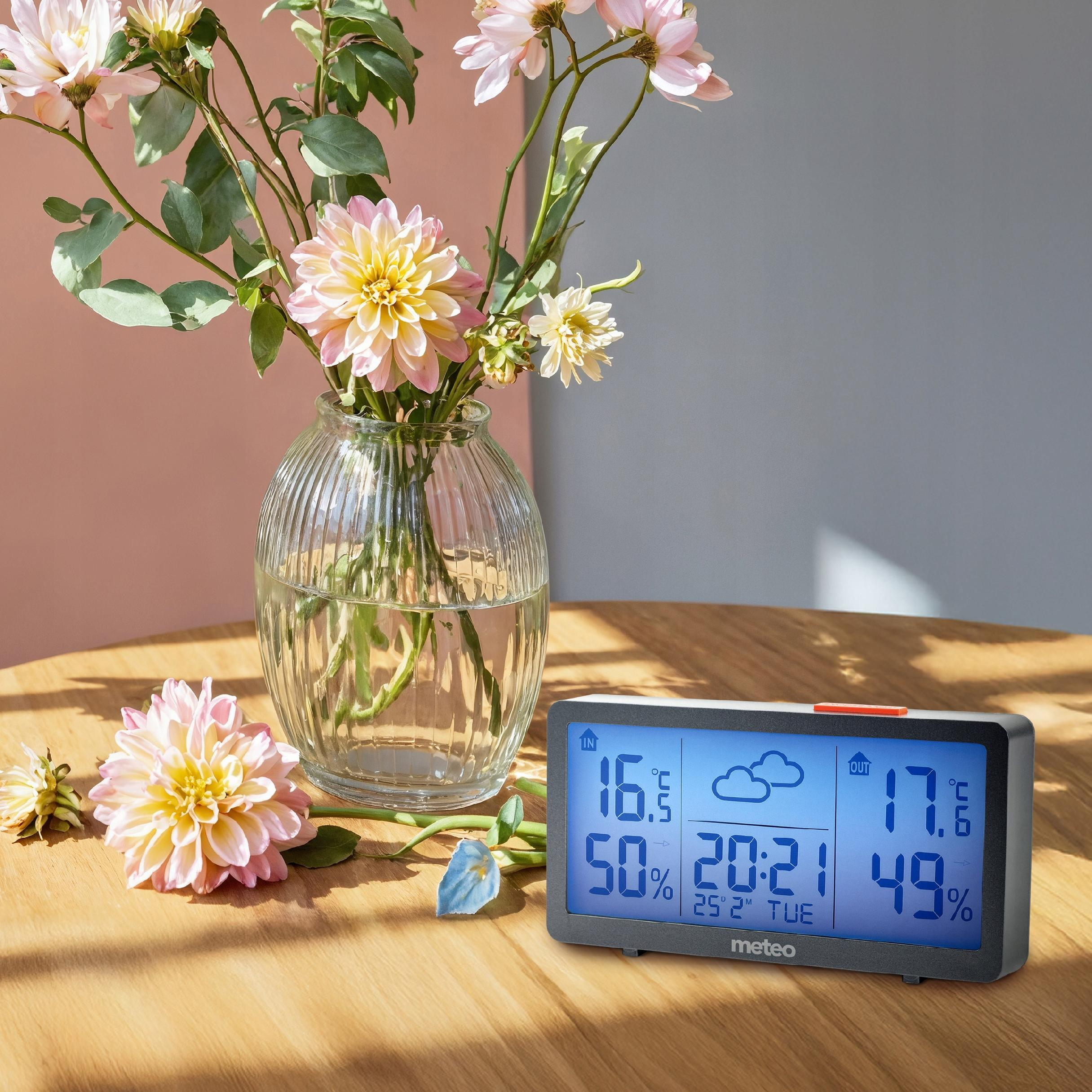 Stół z kwiatami w wazonie i stacja pogodowa pokazująca temperaturę, wilgotność i czas.