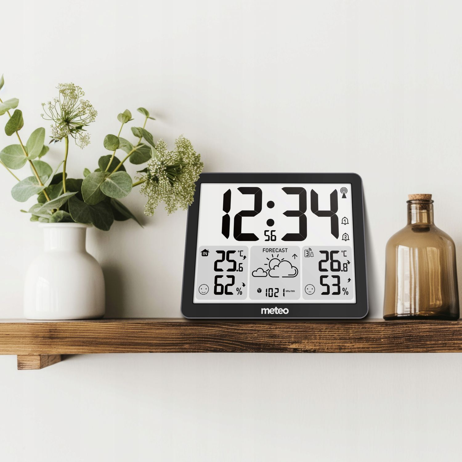 Cyfrowa stacja pogodowa wyświetla godzinę, temperaturę i prognozę pogody na drewnianej półce. Obok znajduje się wazon z roślinami i brązowa butelka.