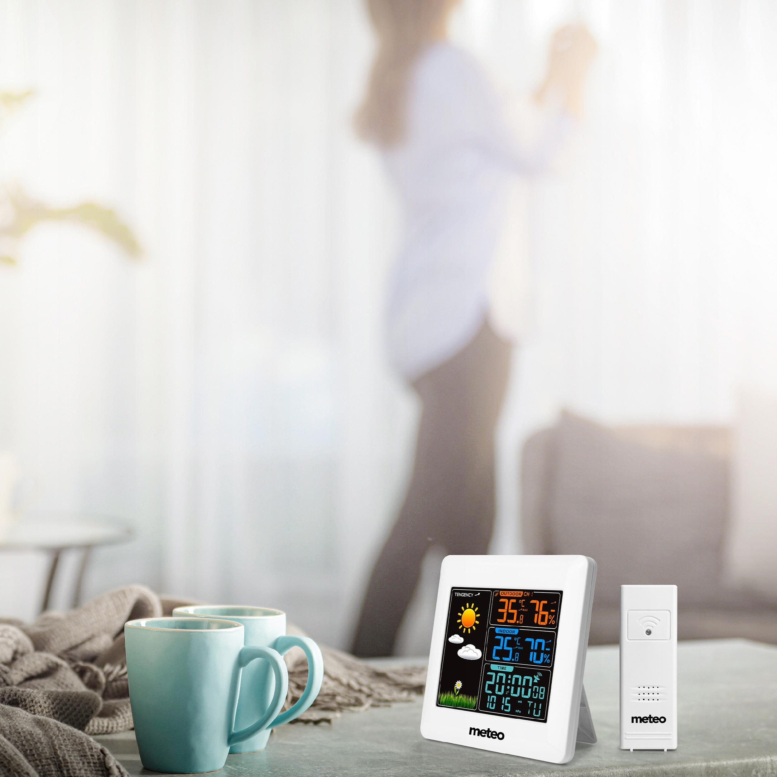 Pokój ze stacją pogodową, kubkami i niewyraźną osobą. Stacja wyświetla temperaturę i godzinę.