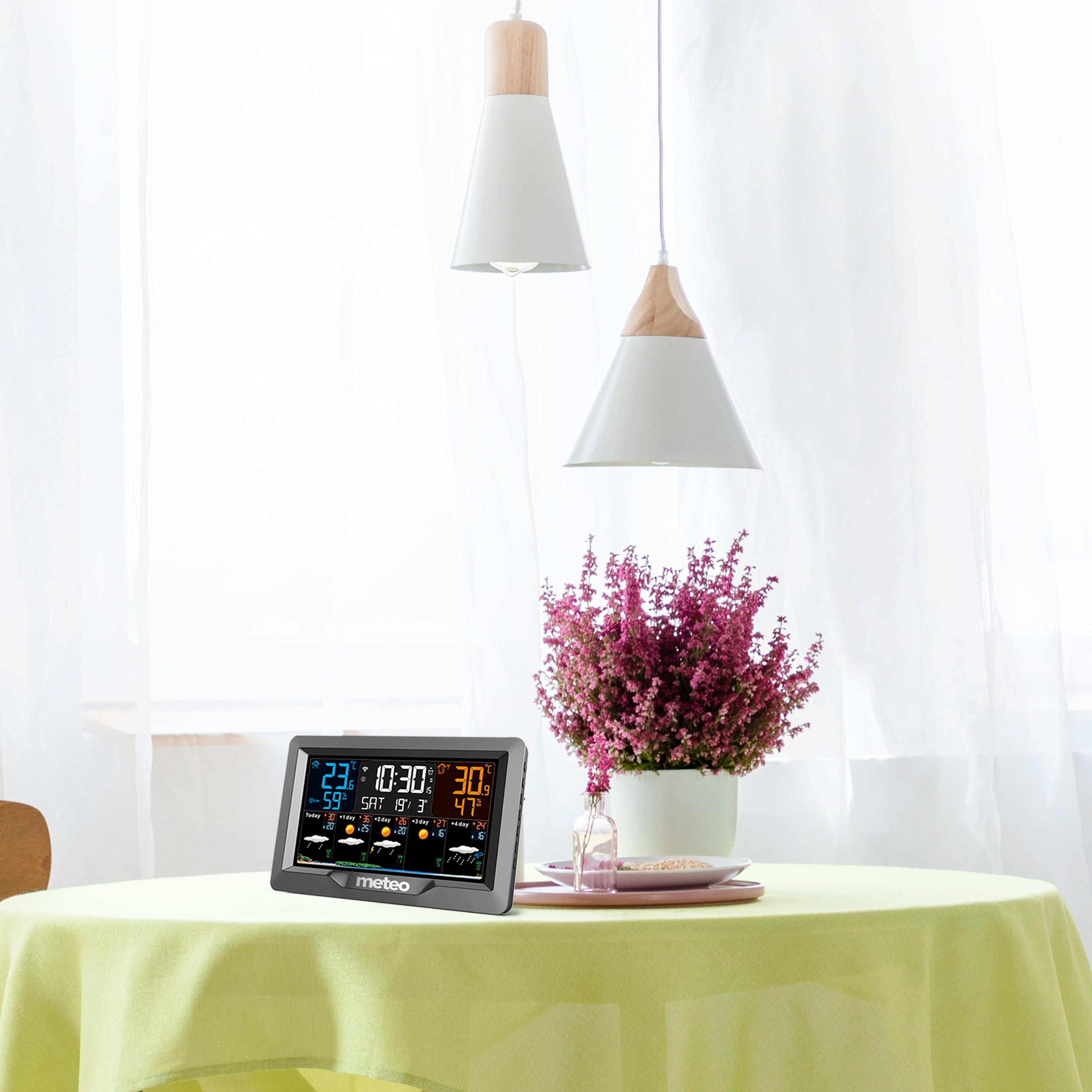 Stół ze stacją pogodową, kwiatami i lampami. Stół ma zielony obrus.