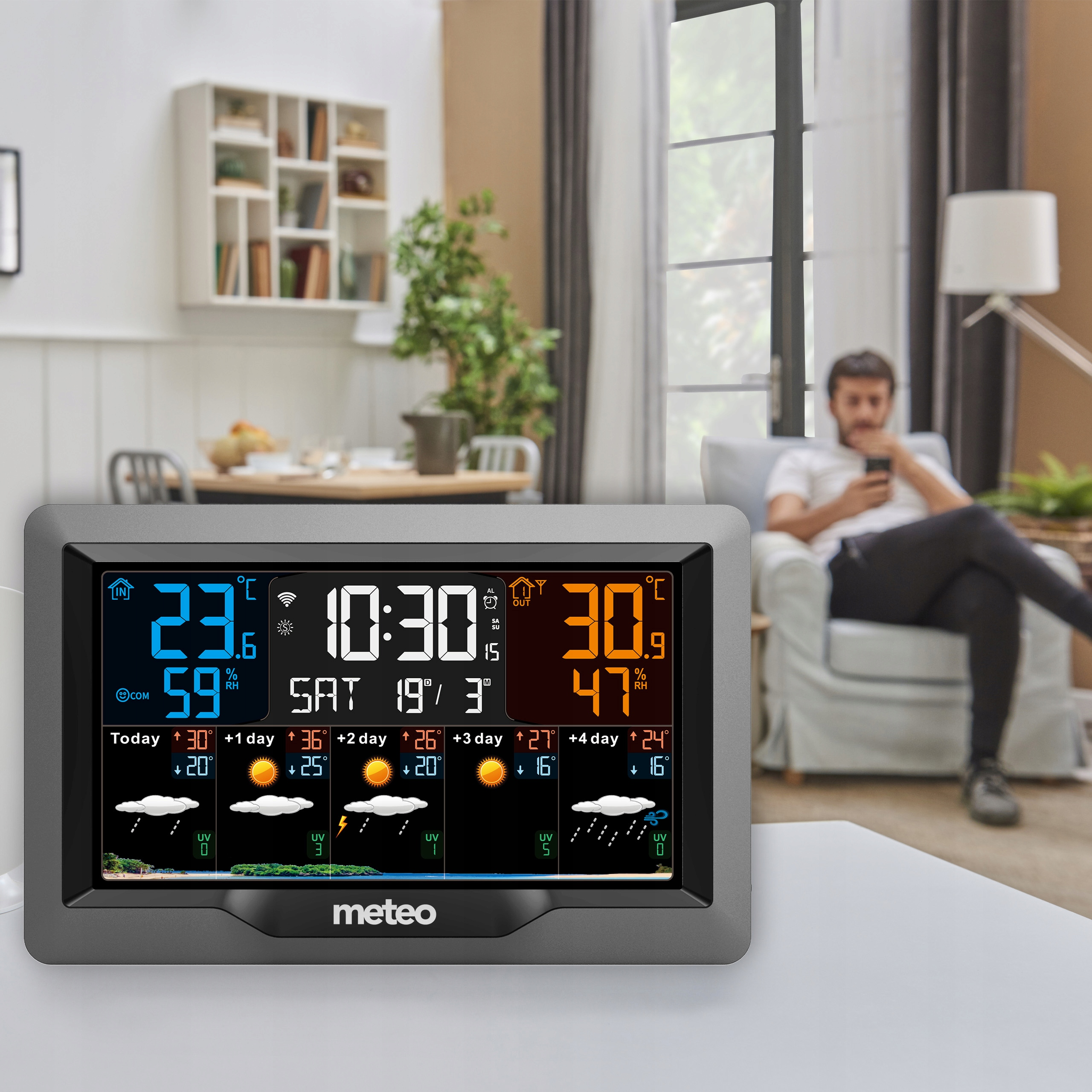 Stacja pogodowa wyświetla temperaturę, wilgotność i prognozę. Osoba siedzi na krześle, korzystając z telefonu w tle.